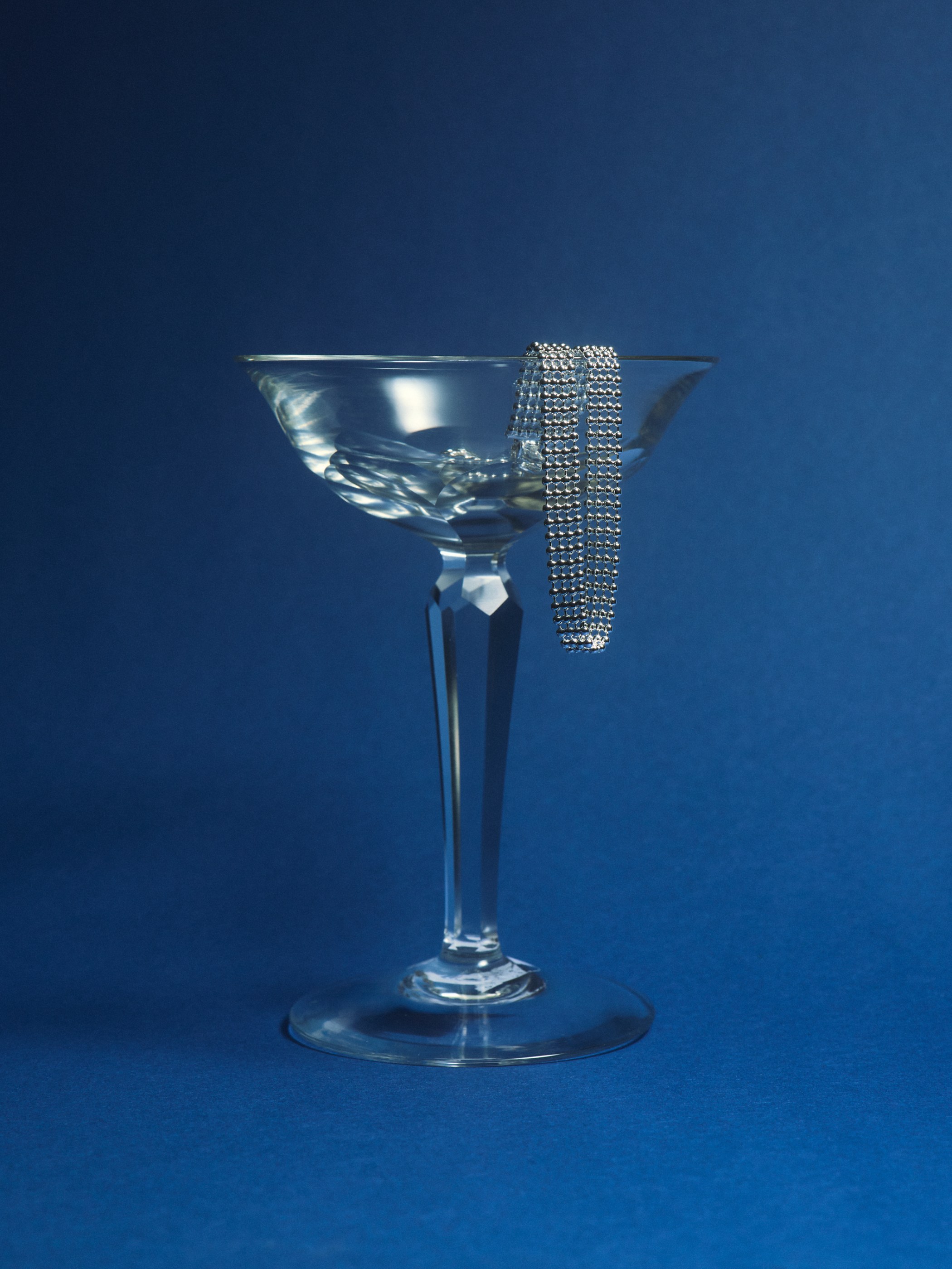 Silver beaded bracelet draped over a clear coupe cocktail glass on a blue background.
