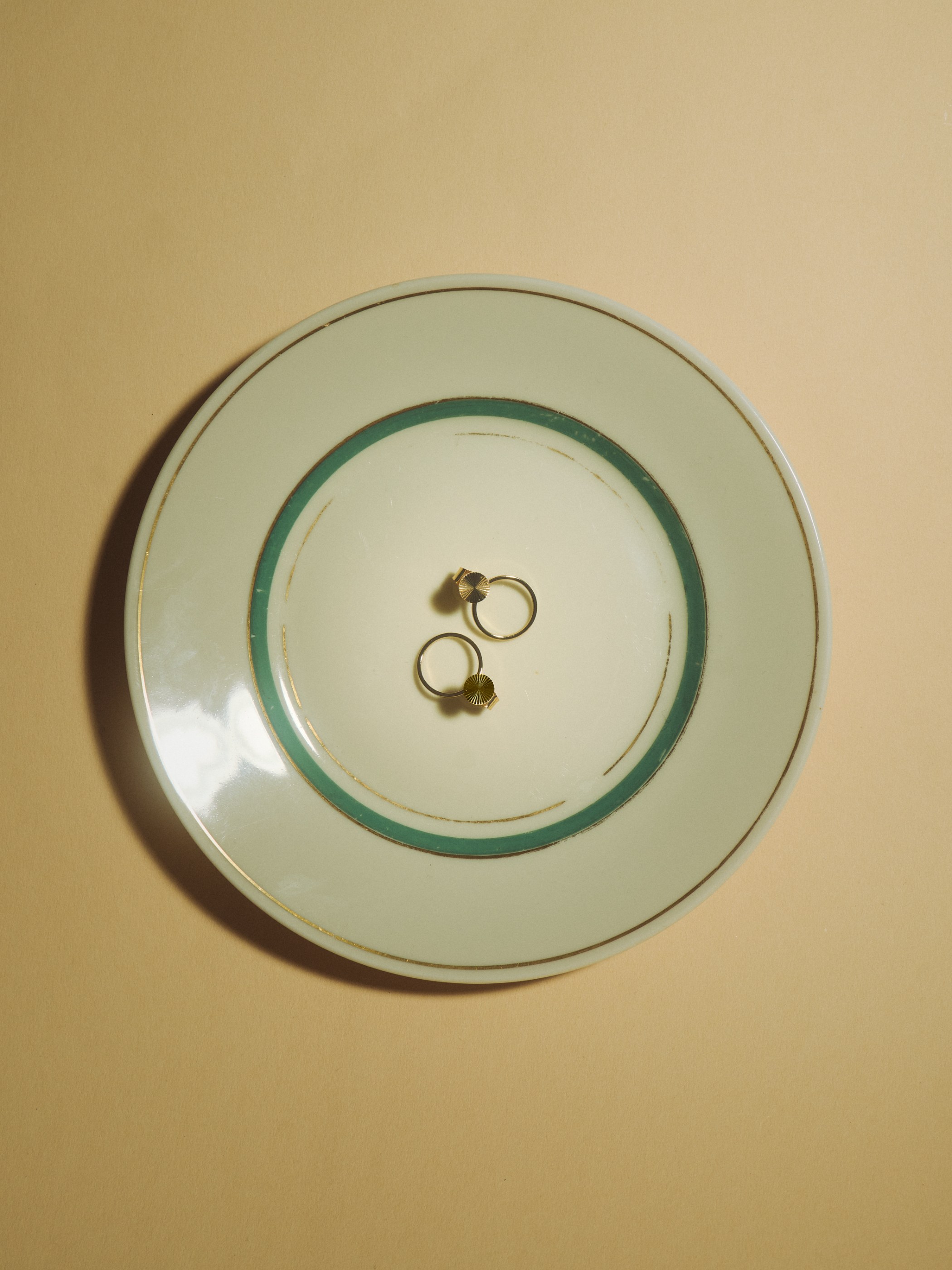 Two small gold earrings on a light green plate with teal and gold bands on a beige surface.