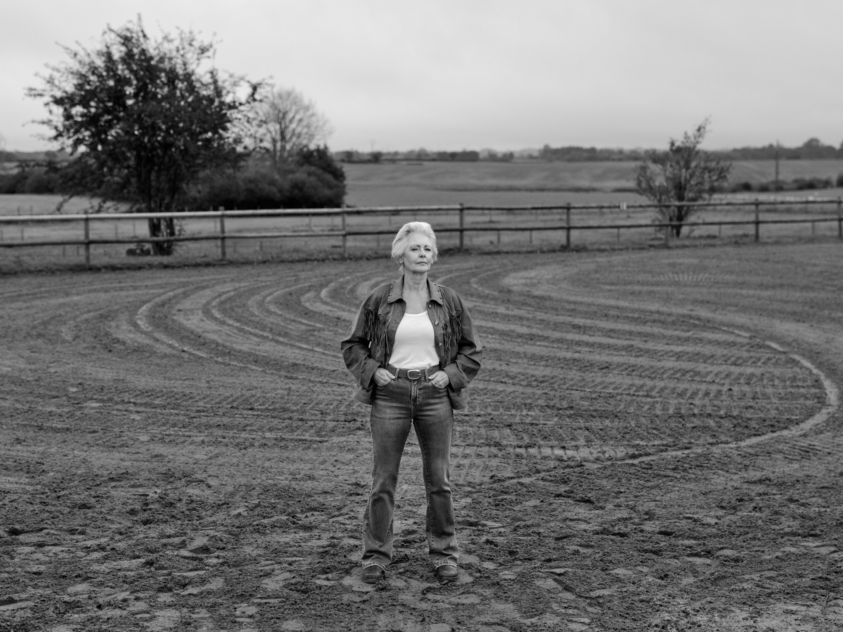 Older woman in a fringed denim jacket and jeans stands in a black and white dirt arena.