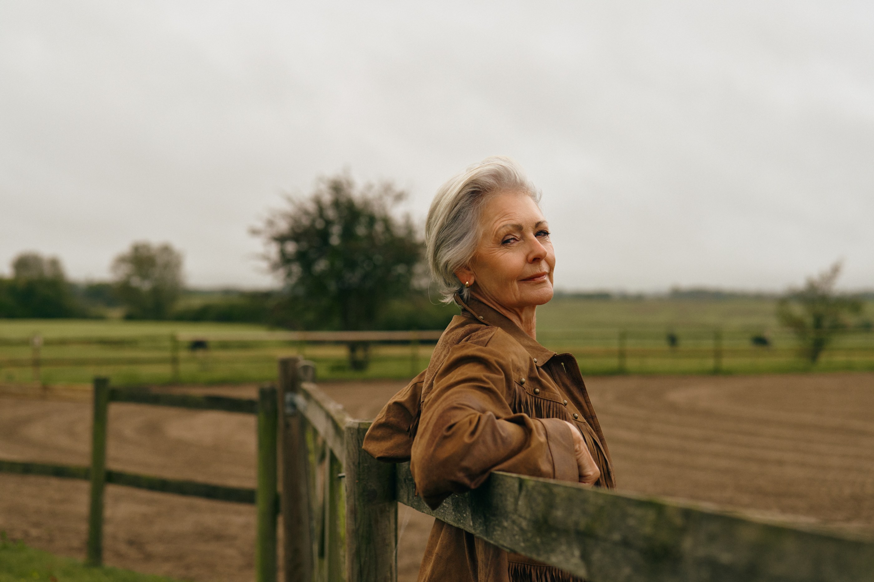 Senior woman in brown fringed jacket looking over her shoulder by a wooden fence.