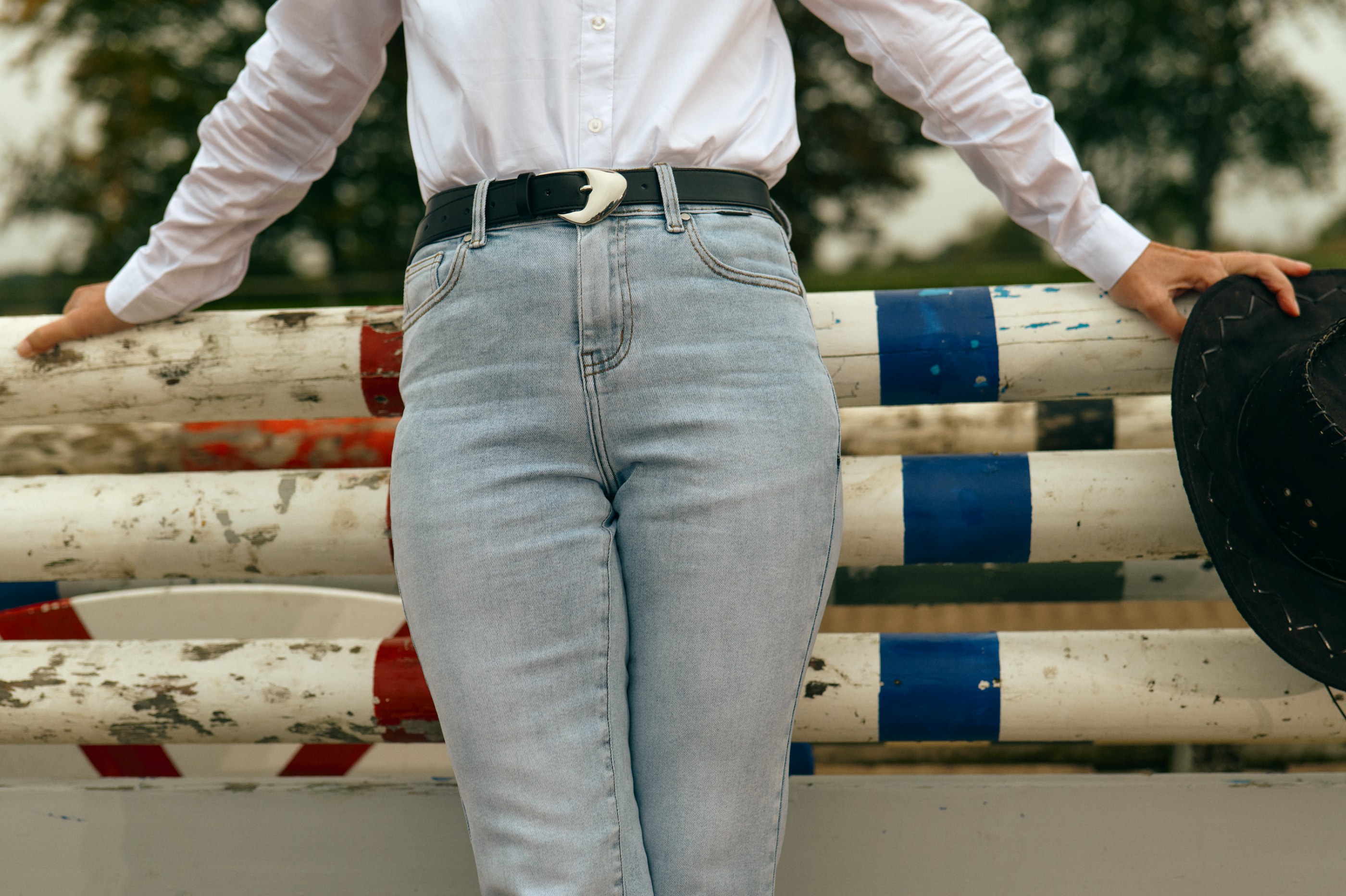 Person in white shirt and light blue jeans leaning on equestrian rails, holding a black hat.