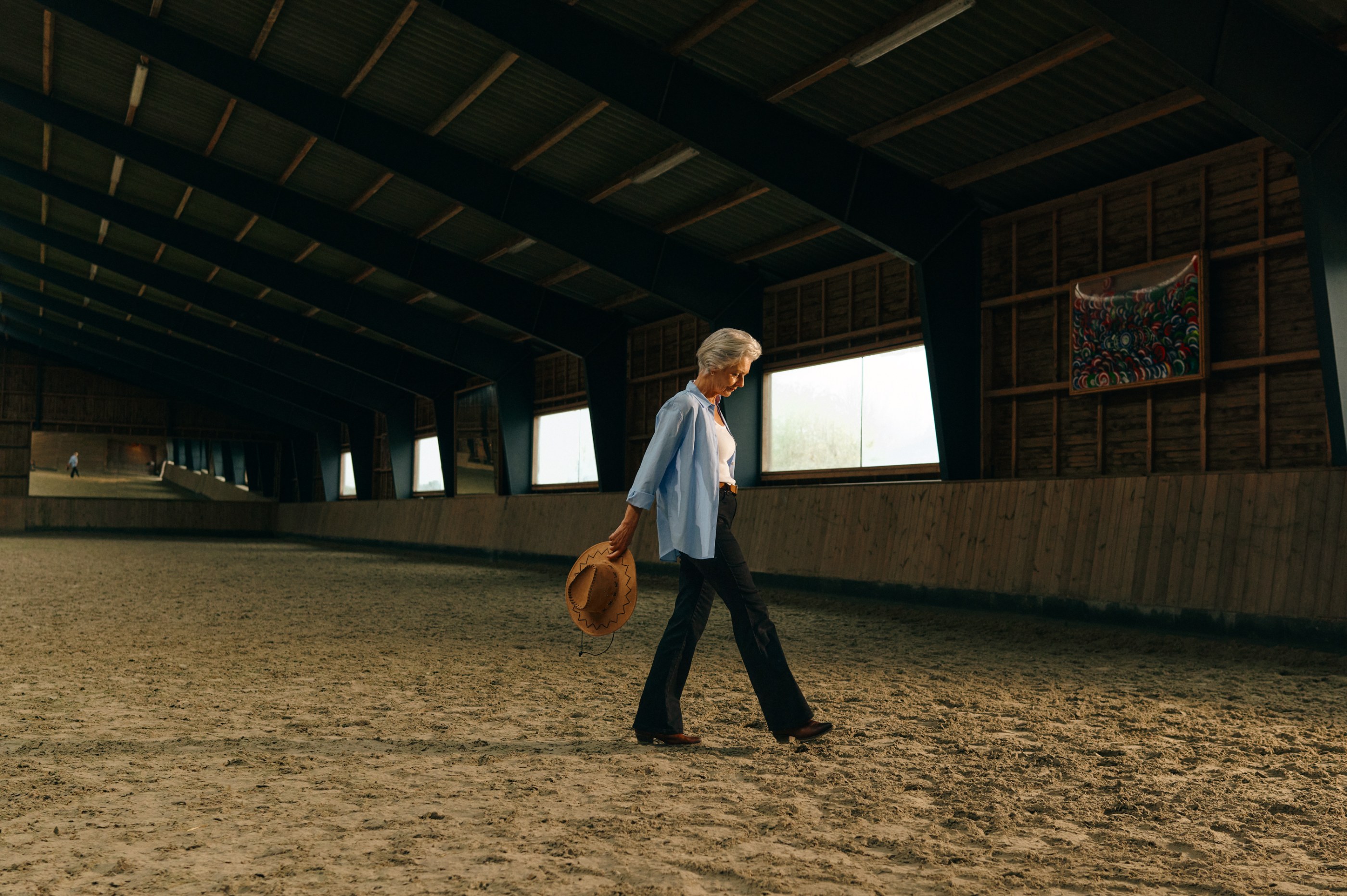 Elegant senior woman walks across a sandy arena, holding a brown cowboy hat.