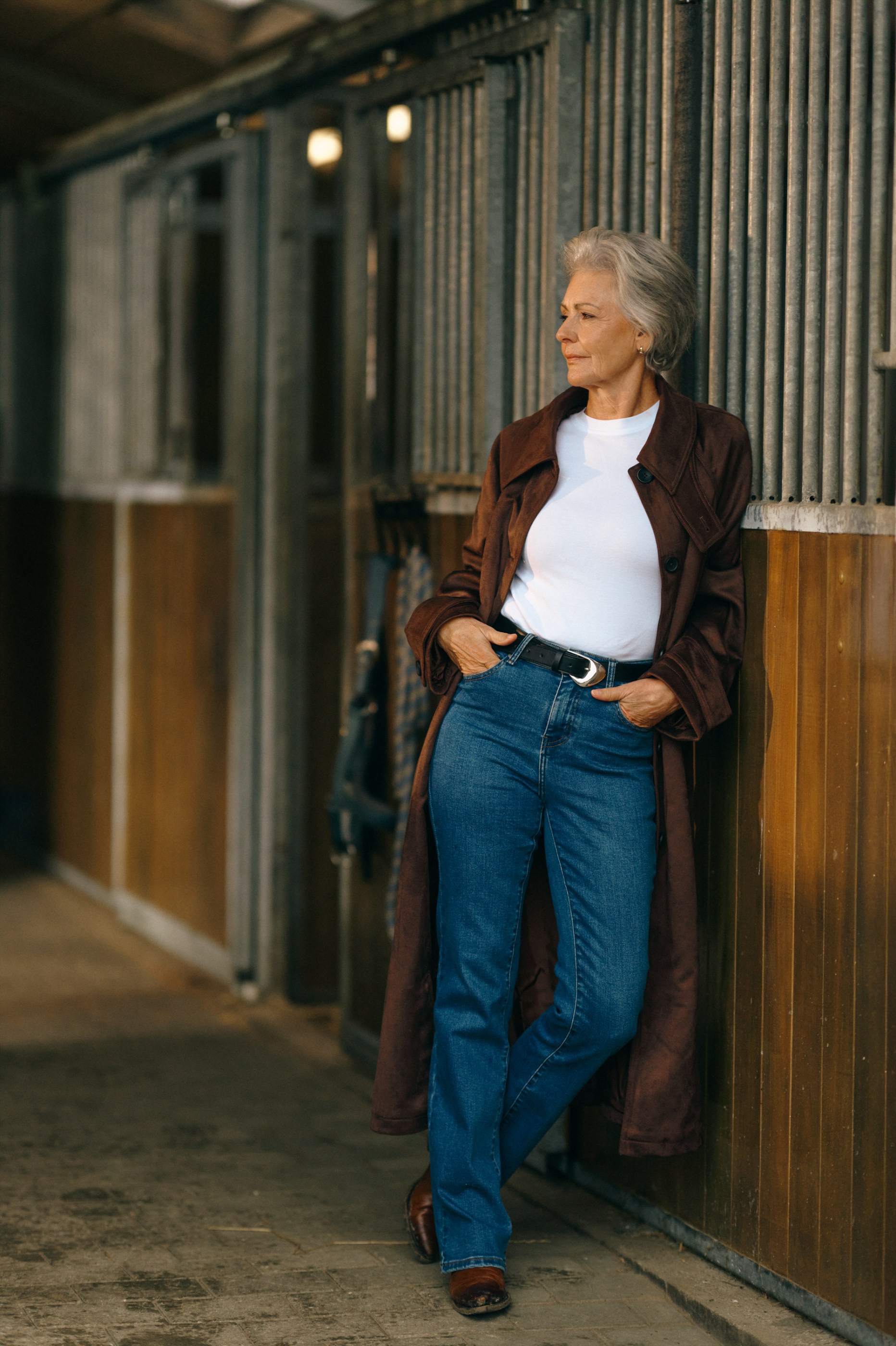 Older woman in a brown coat, white shirt, and blue jeans, leaning against a stable.