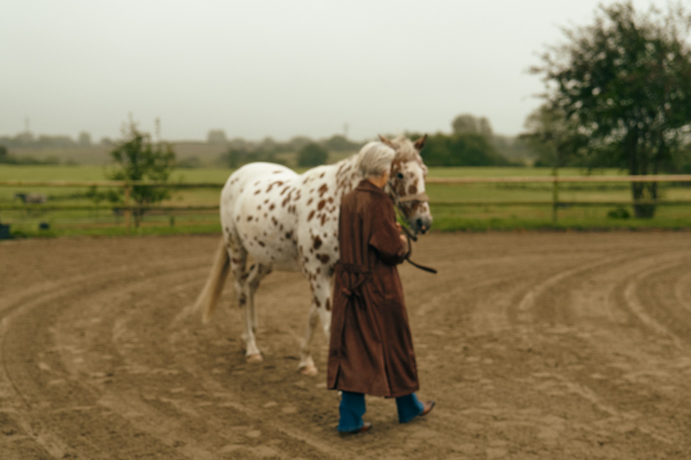 Older person in a brown coat leading a white spotted horse in a dirt arena.