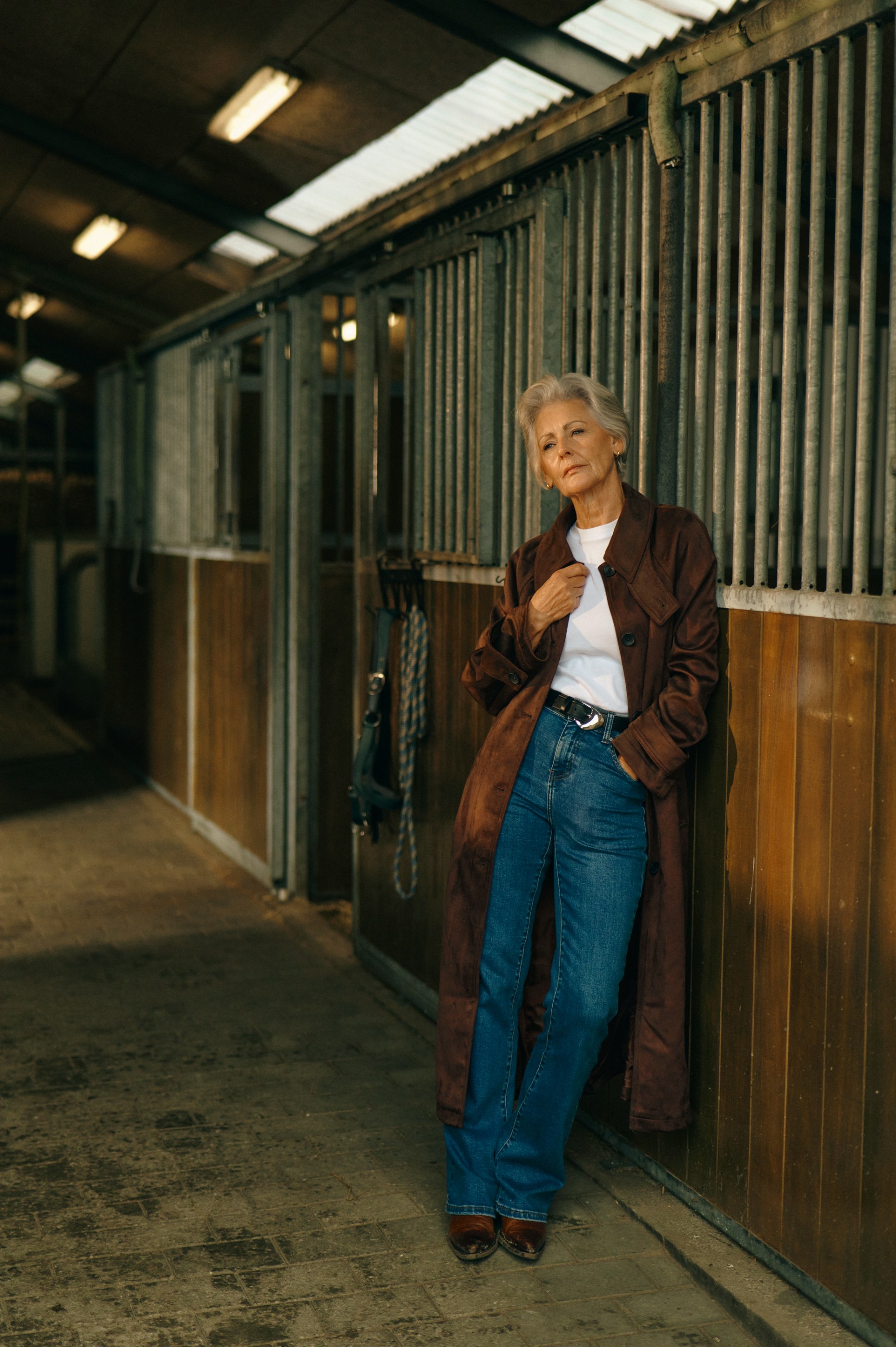 Elderly woman in brown coat and jeans leaning against horse stalls in a stable.