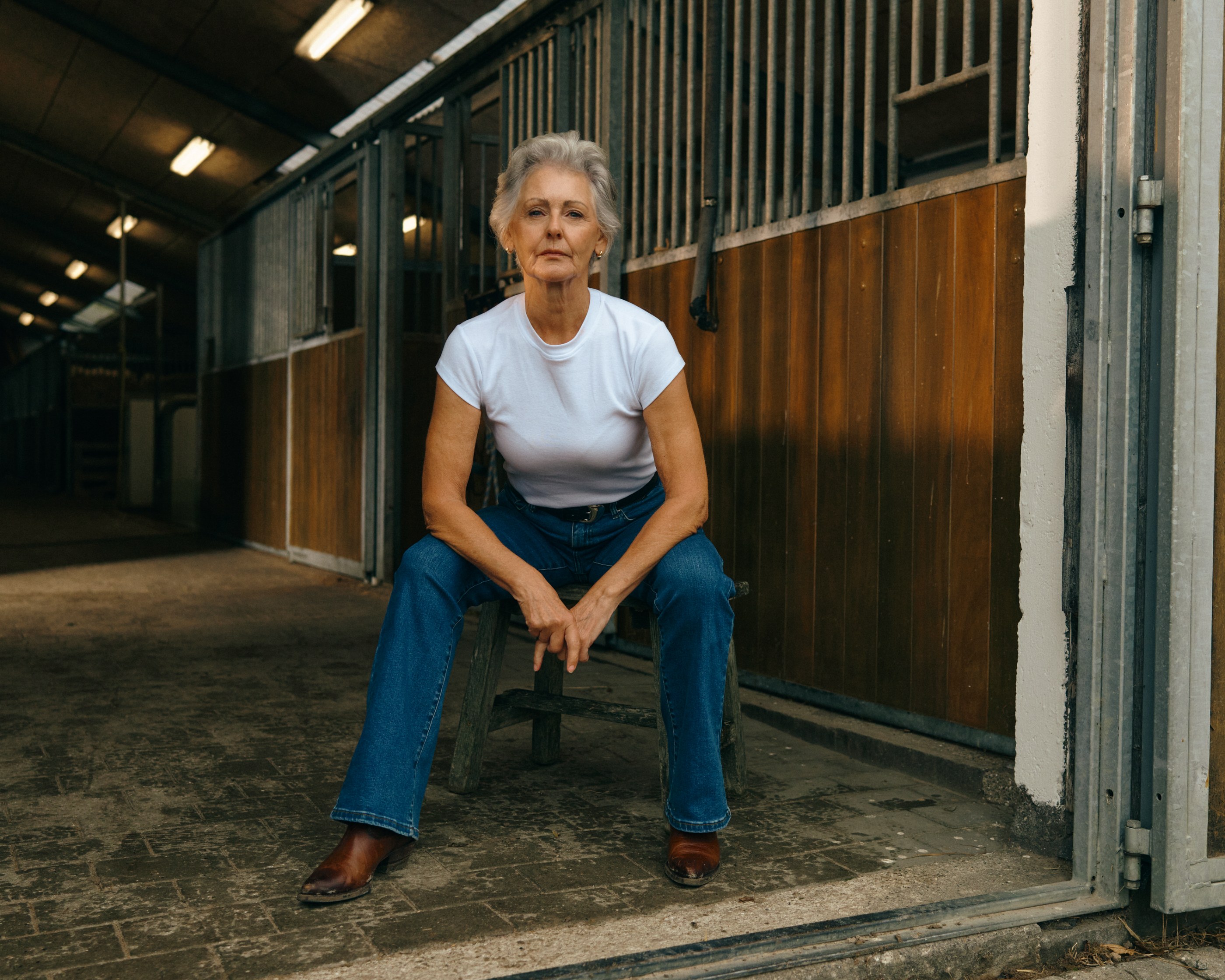 Mature woman with gray hair sits on a stool in a stable, looking at camera.