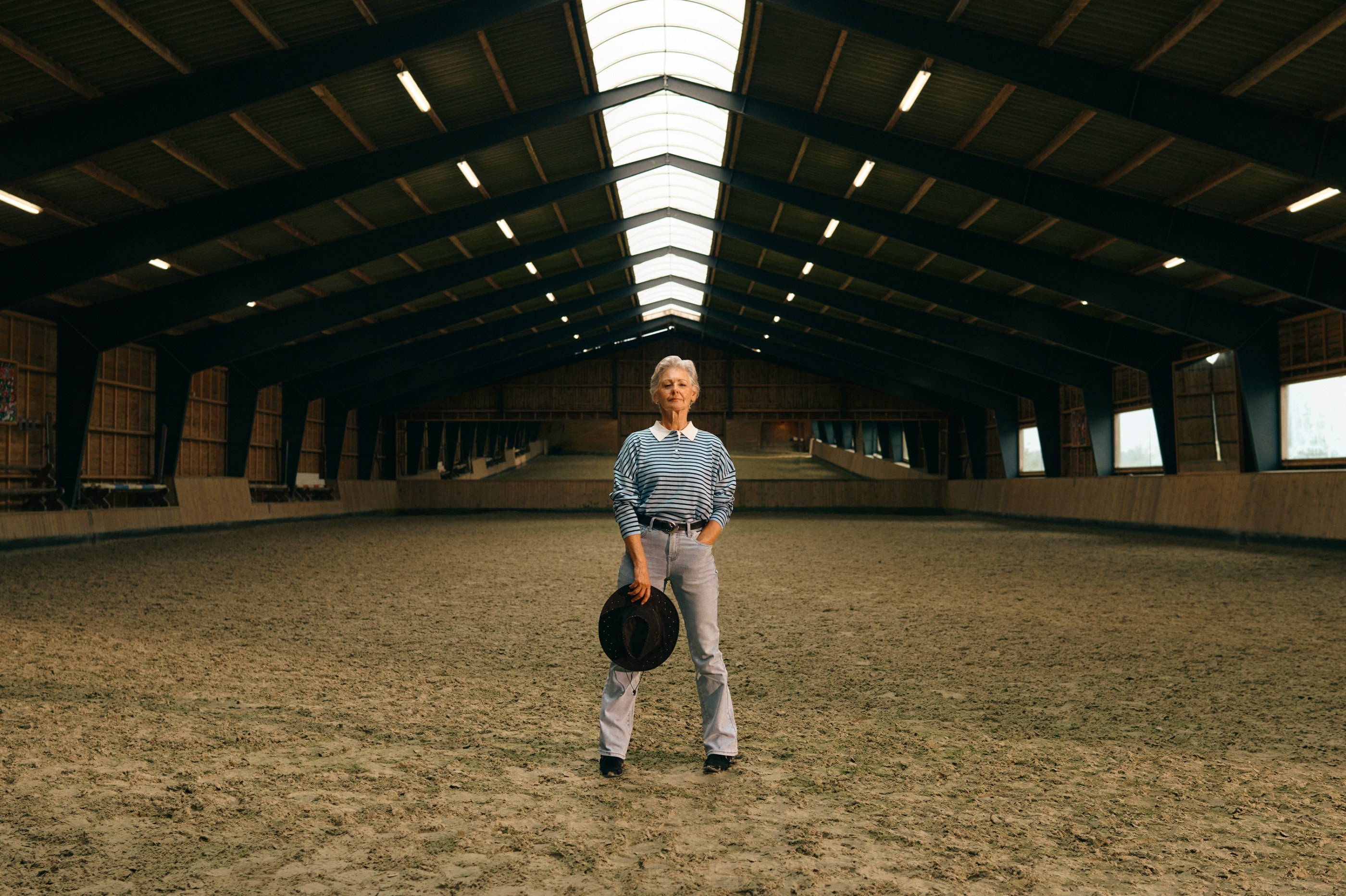 Confident woman holding a hat, standing centered in a well-lit indoor horse arena.