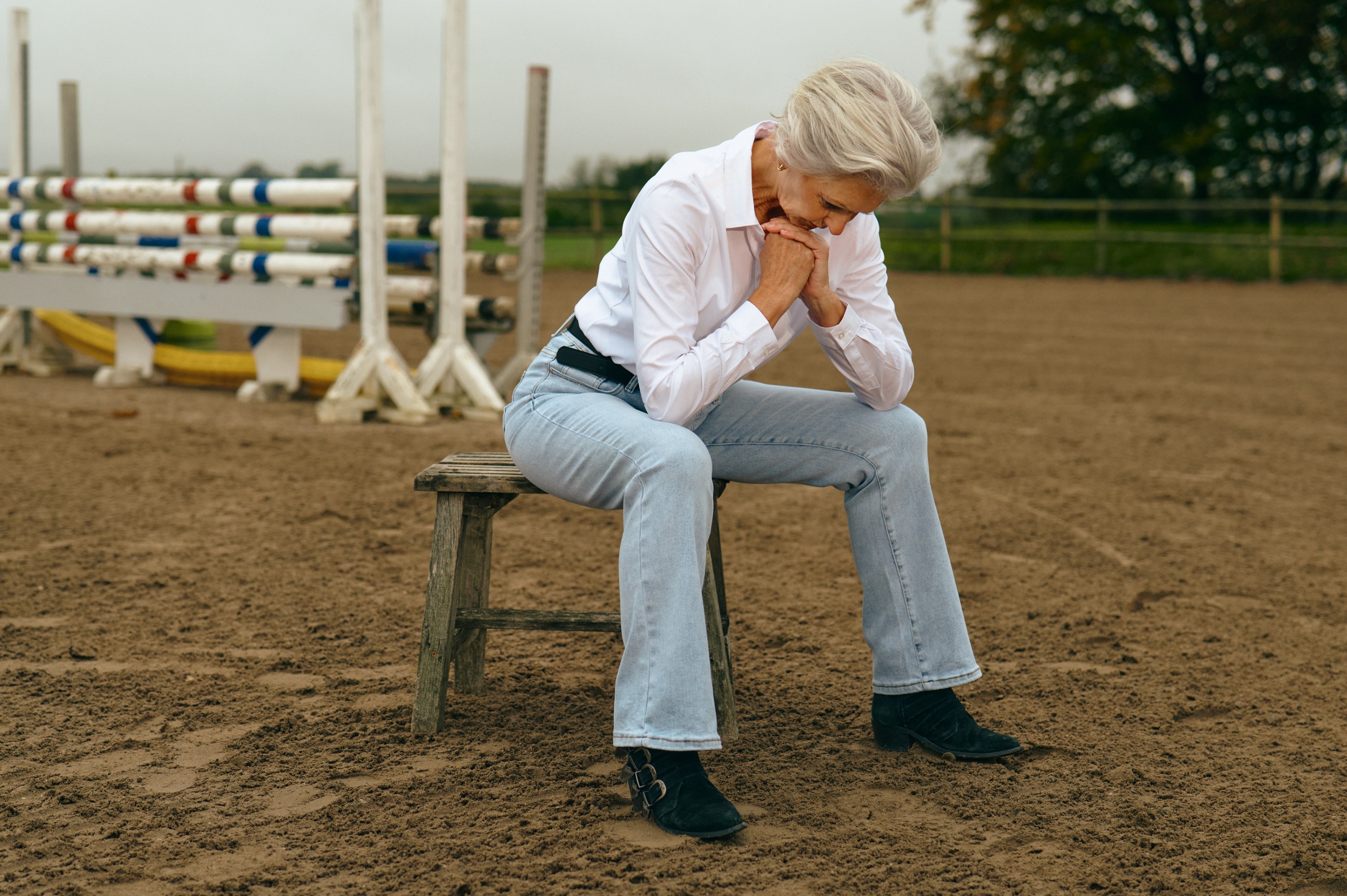 Elderly woman sits on a wooden stool, hands clasped, in an outdoor riding arena.