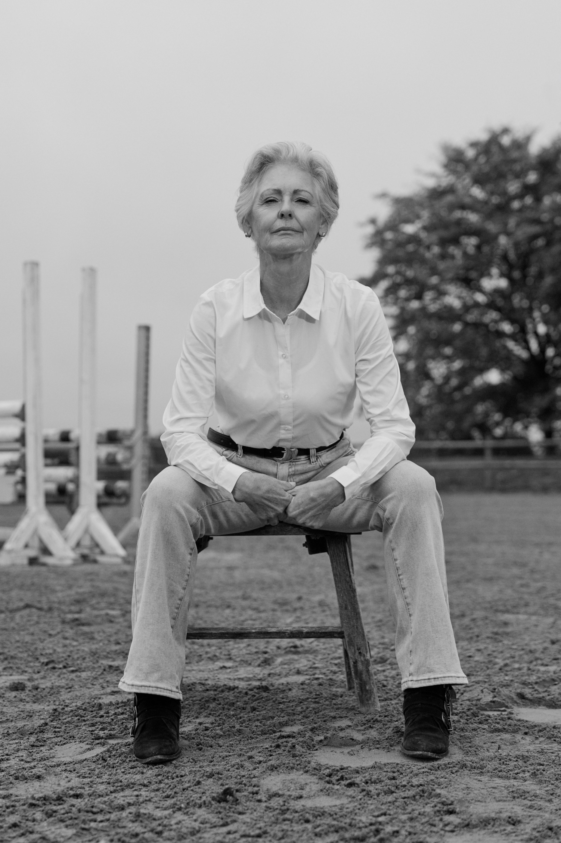 Black and white portrait of an older woman sitting on a stool in an equestrian arena.