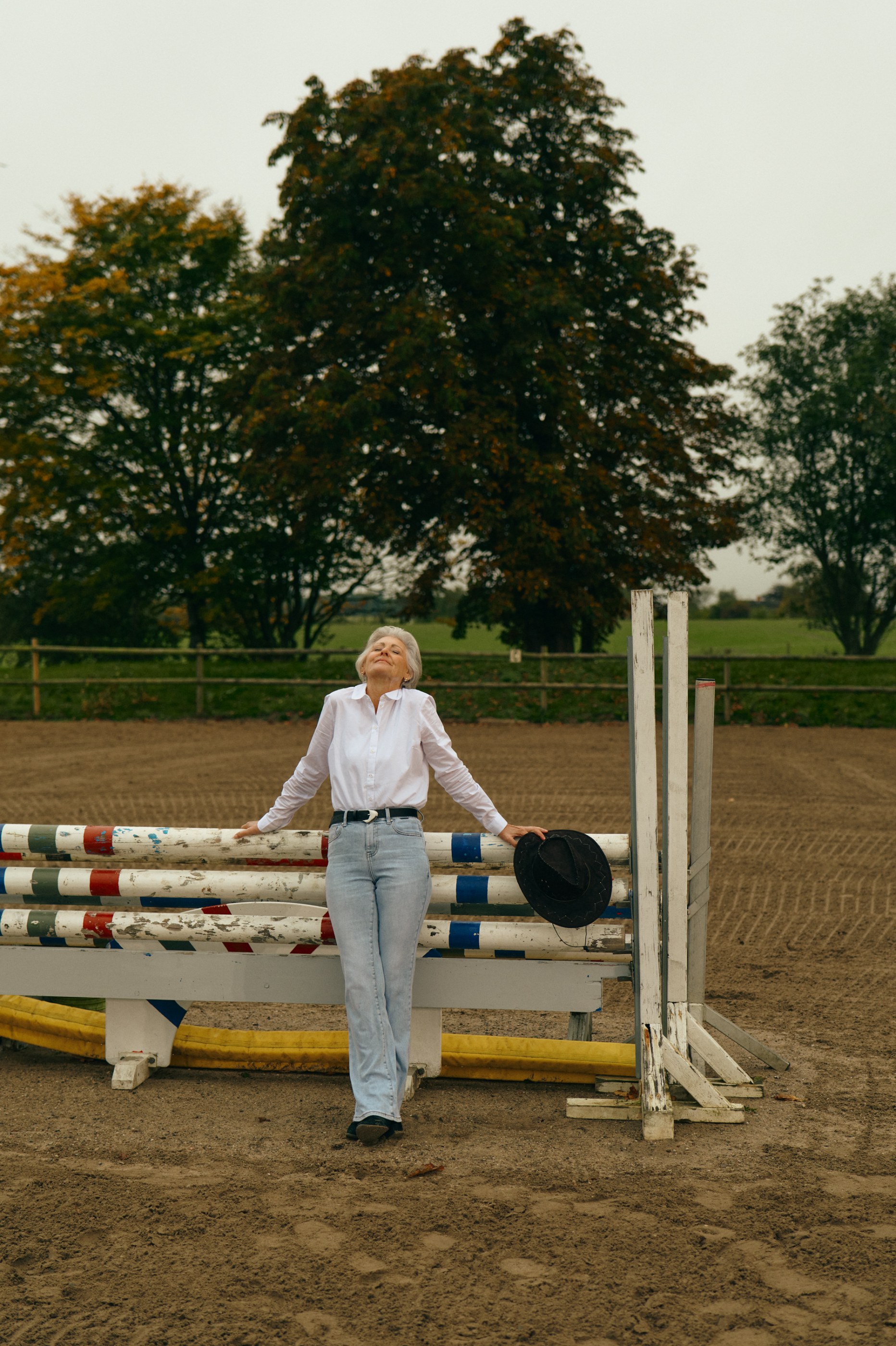 Peaceful senior woman in white shirt and jeans leaning on equestrian jump, holding a hat.