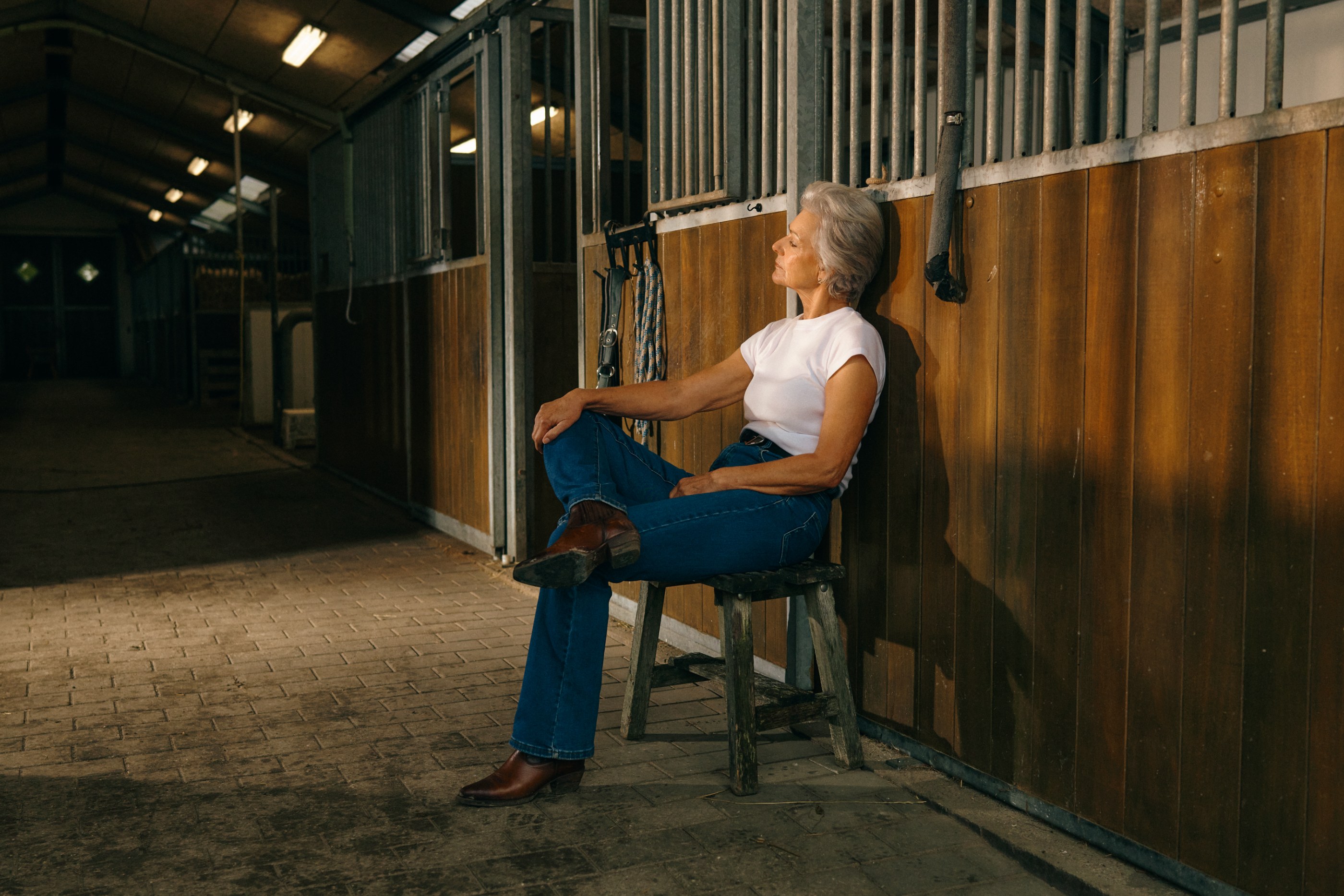 Relaxed older woman in a white t-shirt and jeans sits on a stool in a sunlit stable.