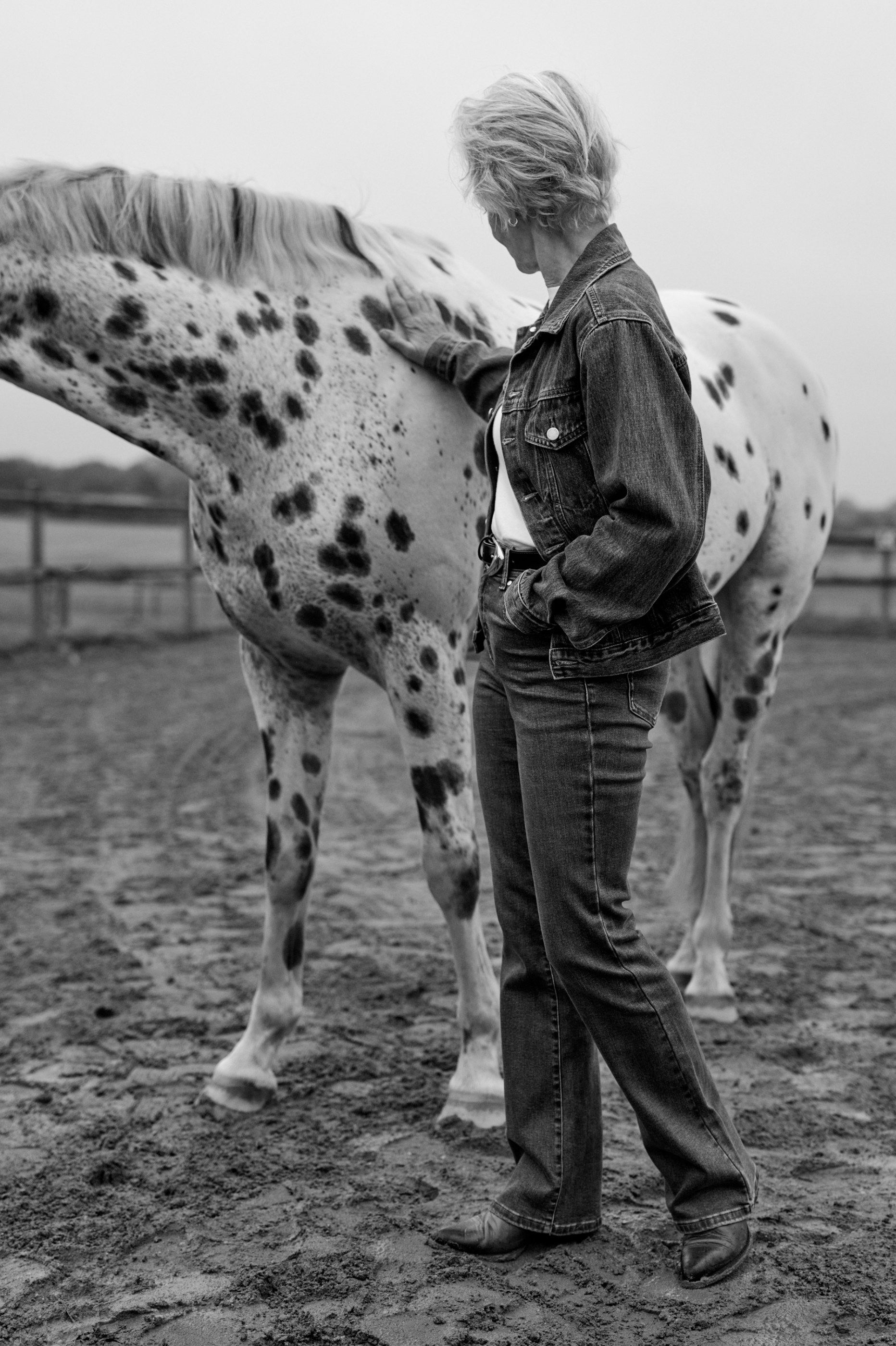Woman in denim jacket and jeans petting a spotted horse, black and white outdoor scene.