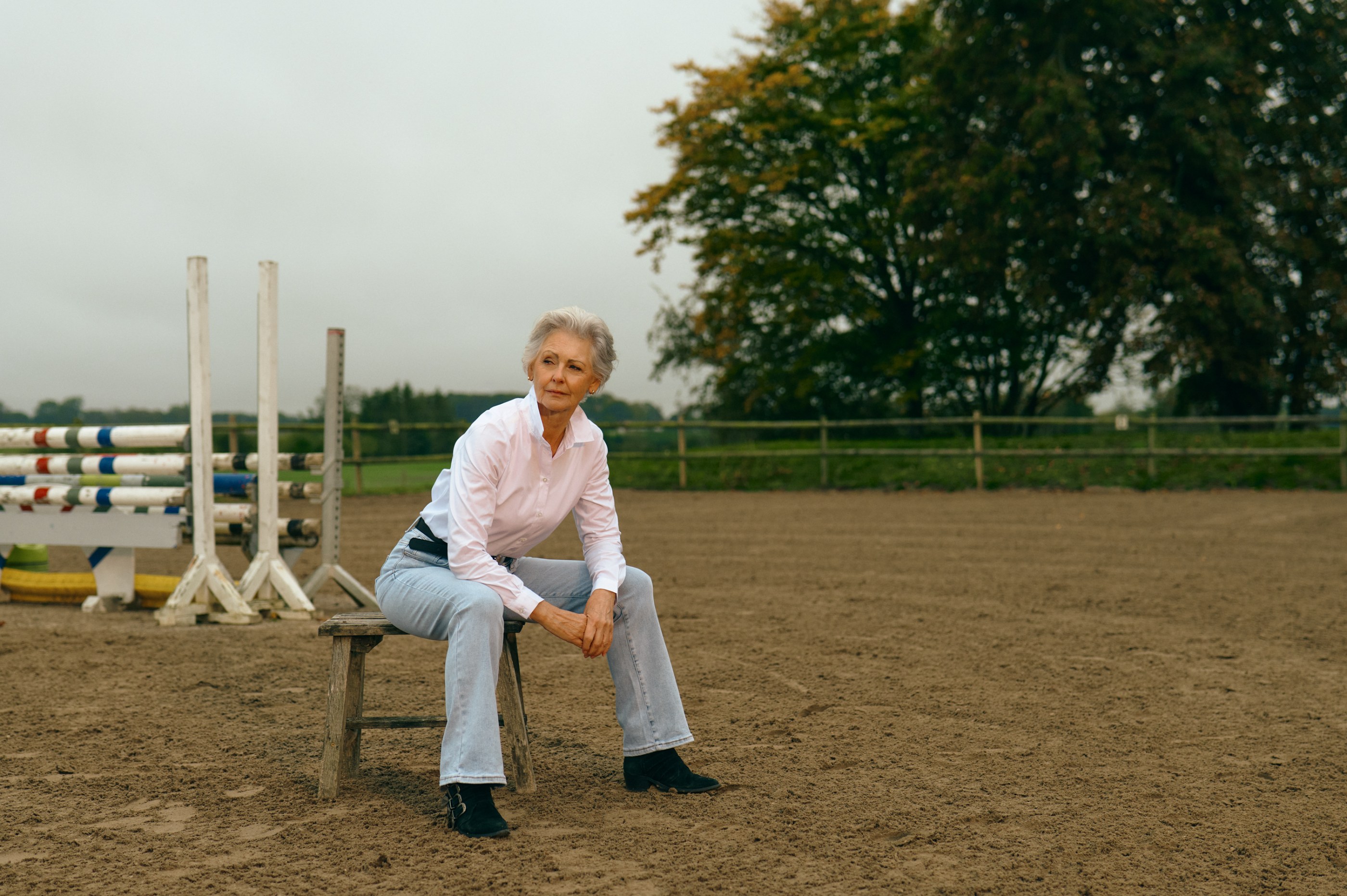 Pensive older woman in white shirt, jeans, and boots sits in a horse arena.