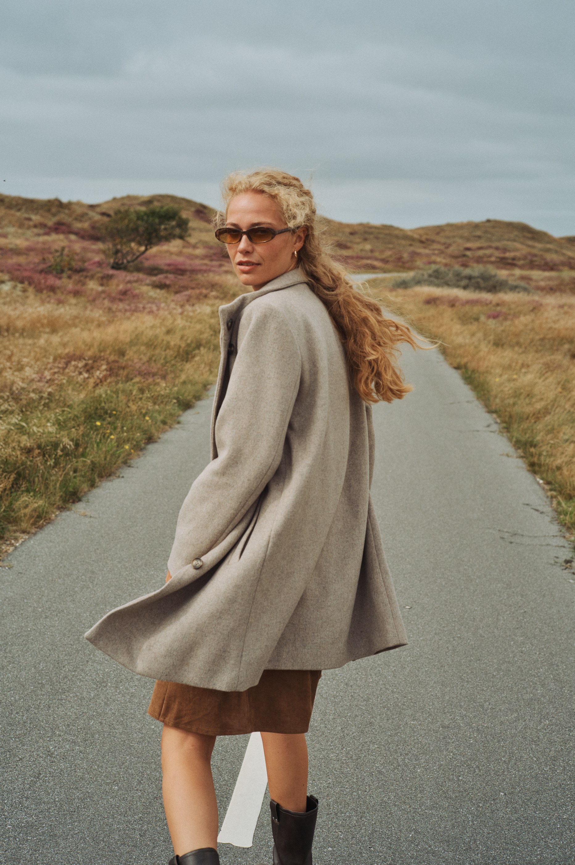 Blonde woman in sunglasses and a beige coat looking back on a road through rolling hills.