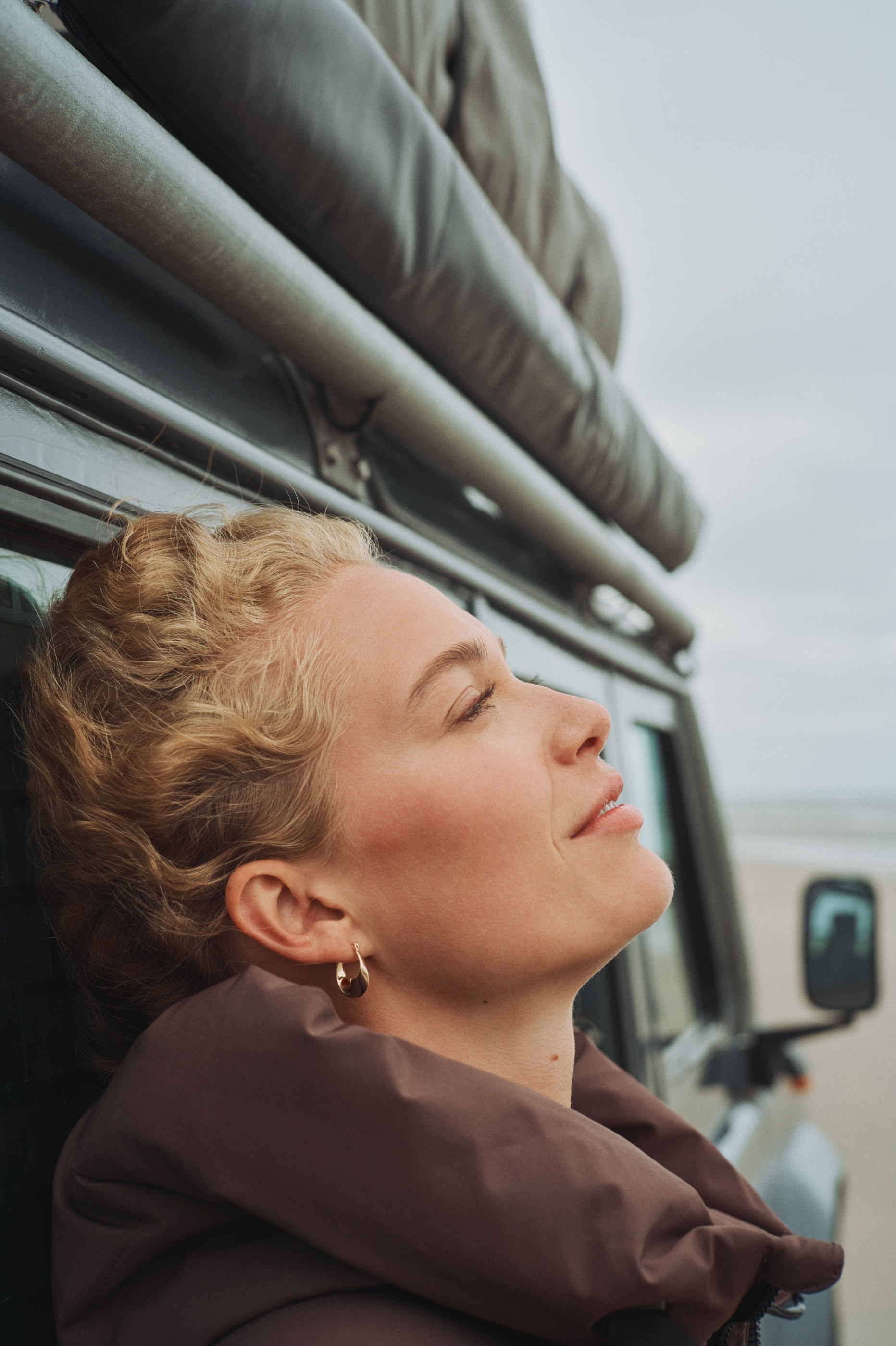 Close-up of a blonde woman with closed eyes, leaning on a vehicle, looking up at the sky.