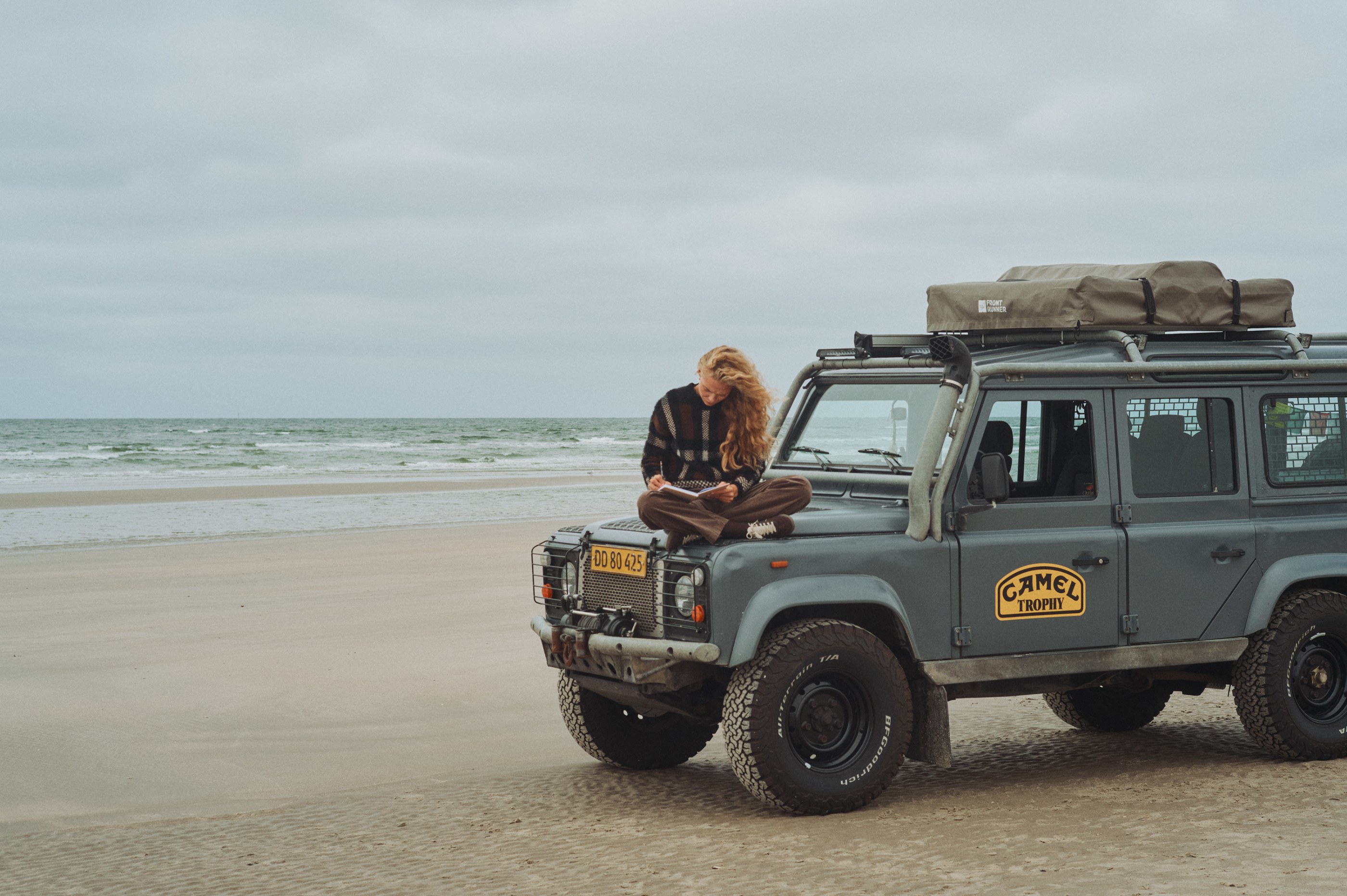 Curly-haired person writing on a gray Land Rover Defender 'Camel Trophy' on a cloudy beach.