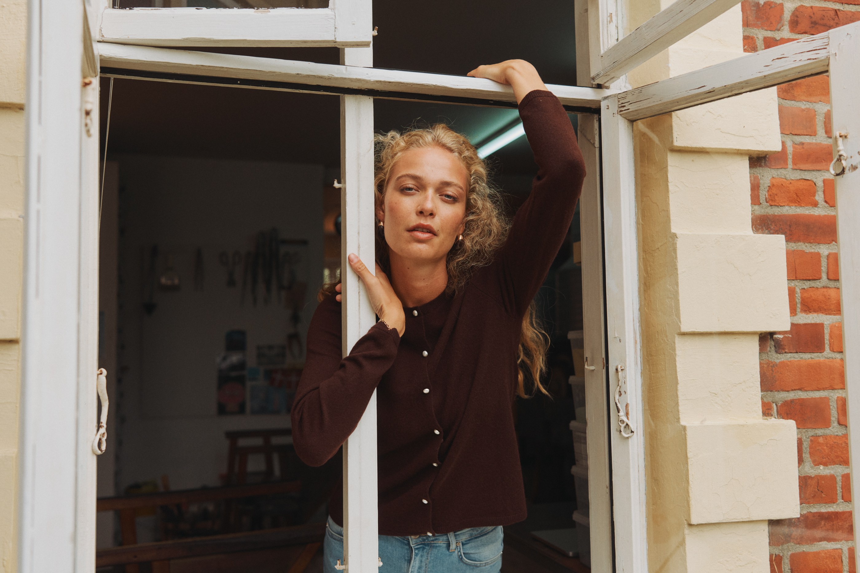 Woman with curly blonde hair in a brown cardigan framed by a rustic white window.