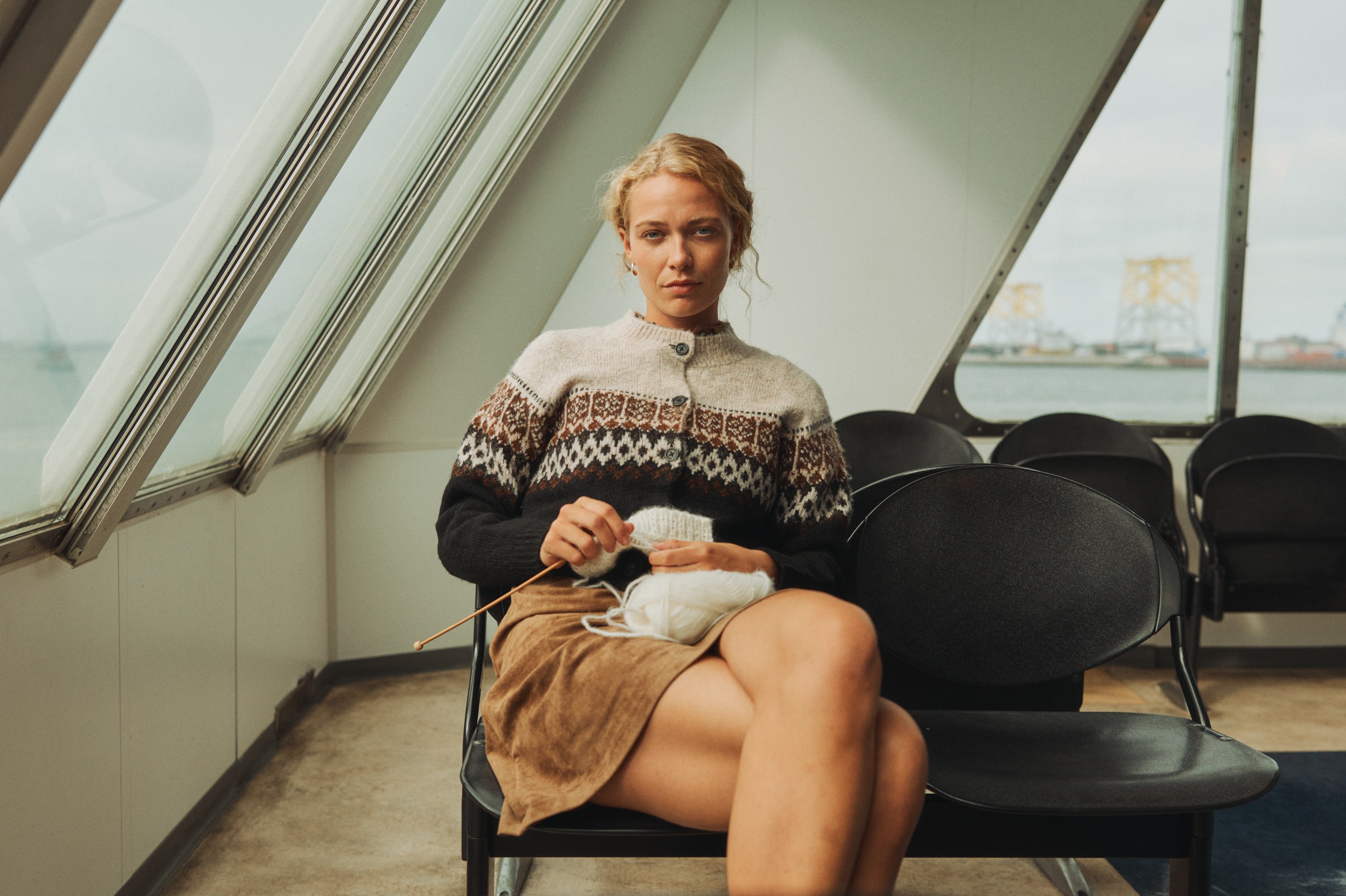 A woman in a patterned cardigan knits on a ferry with an industrial backdrop.