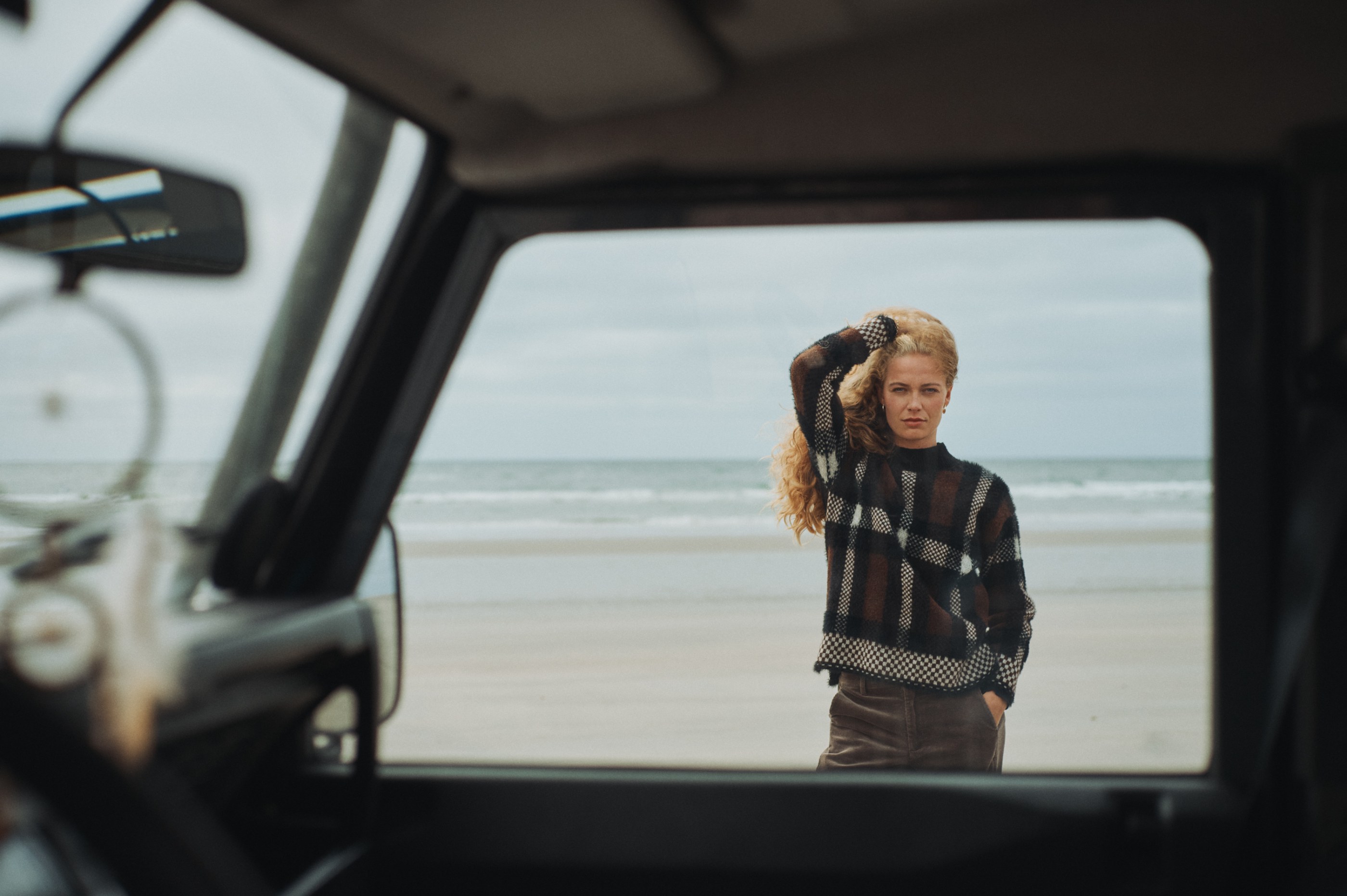 Curly-haired woman in plaid sweater on beach, framed by car window.