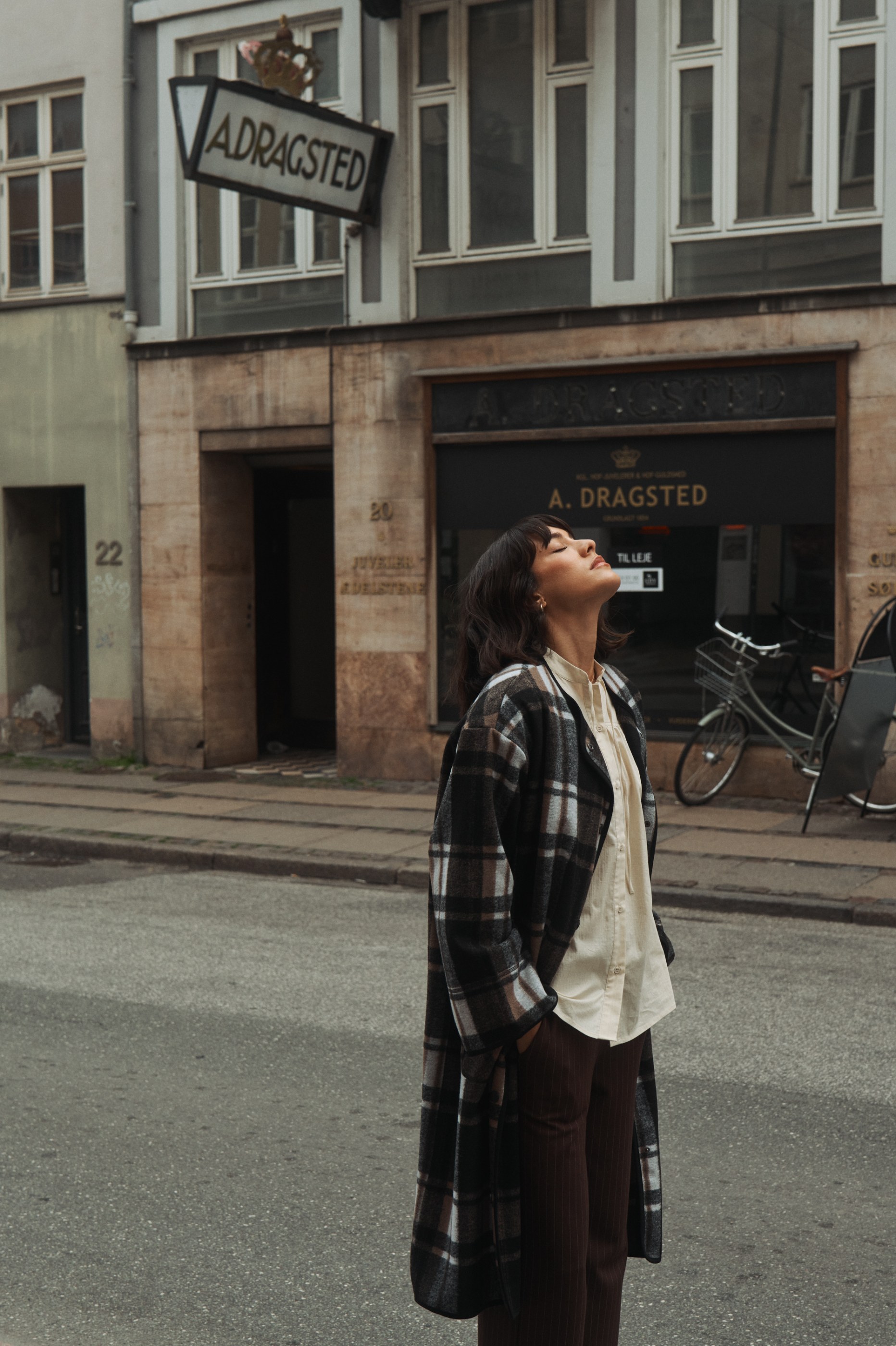 Woman in plaid coat and pinstriped pants looks up on a street with 'ADRAGSTED' signs.