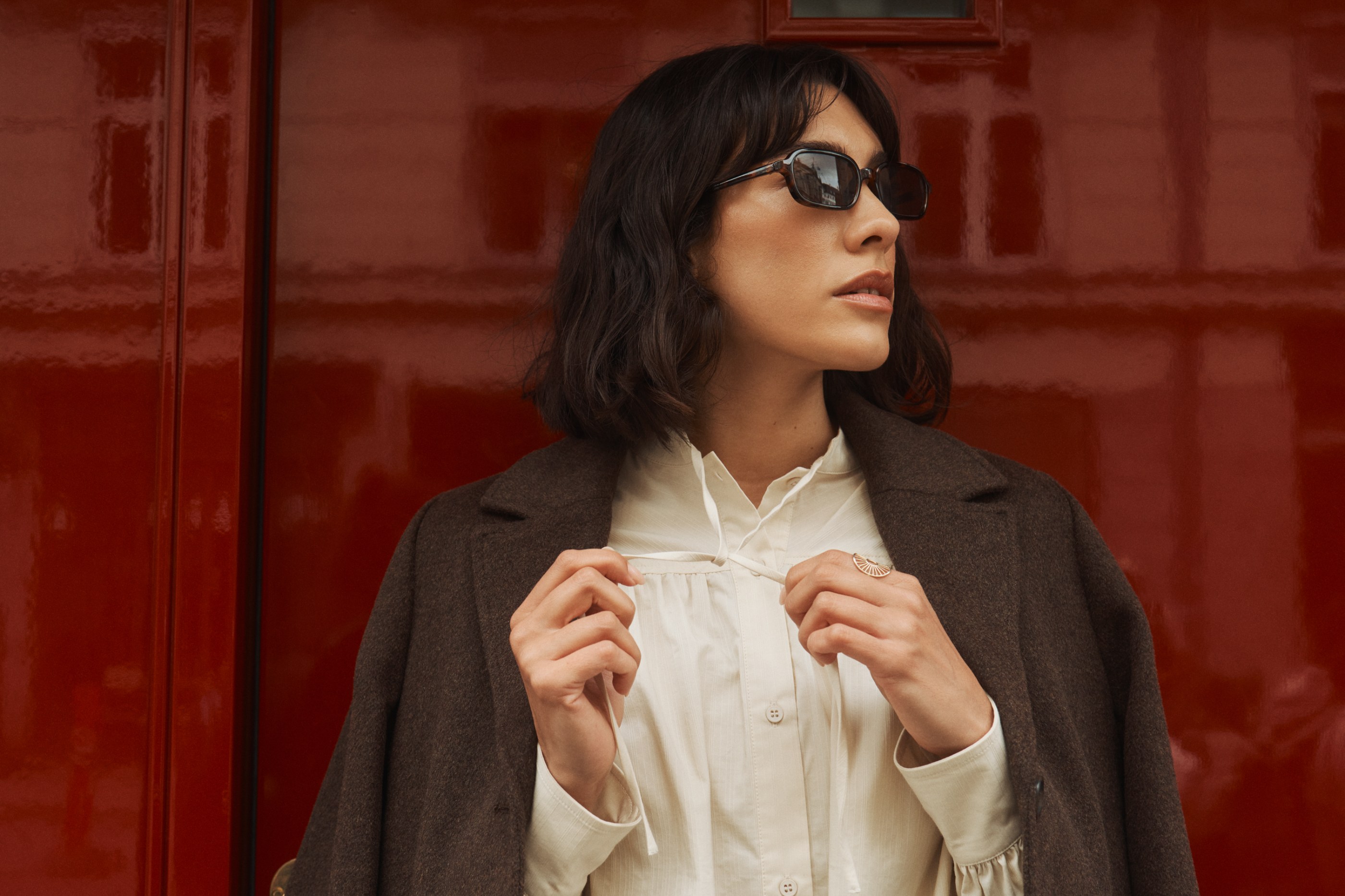Woman in sunglasses and brown coat, holding blouse ties, against a glossy red background.