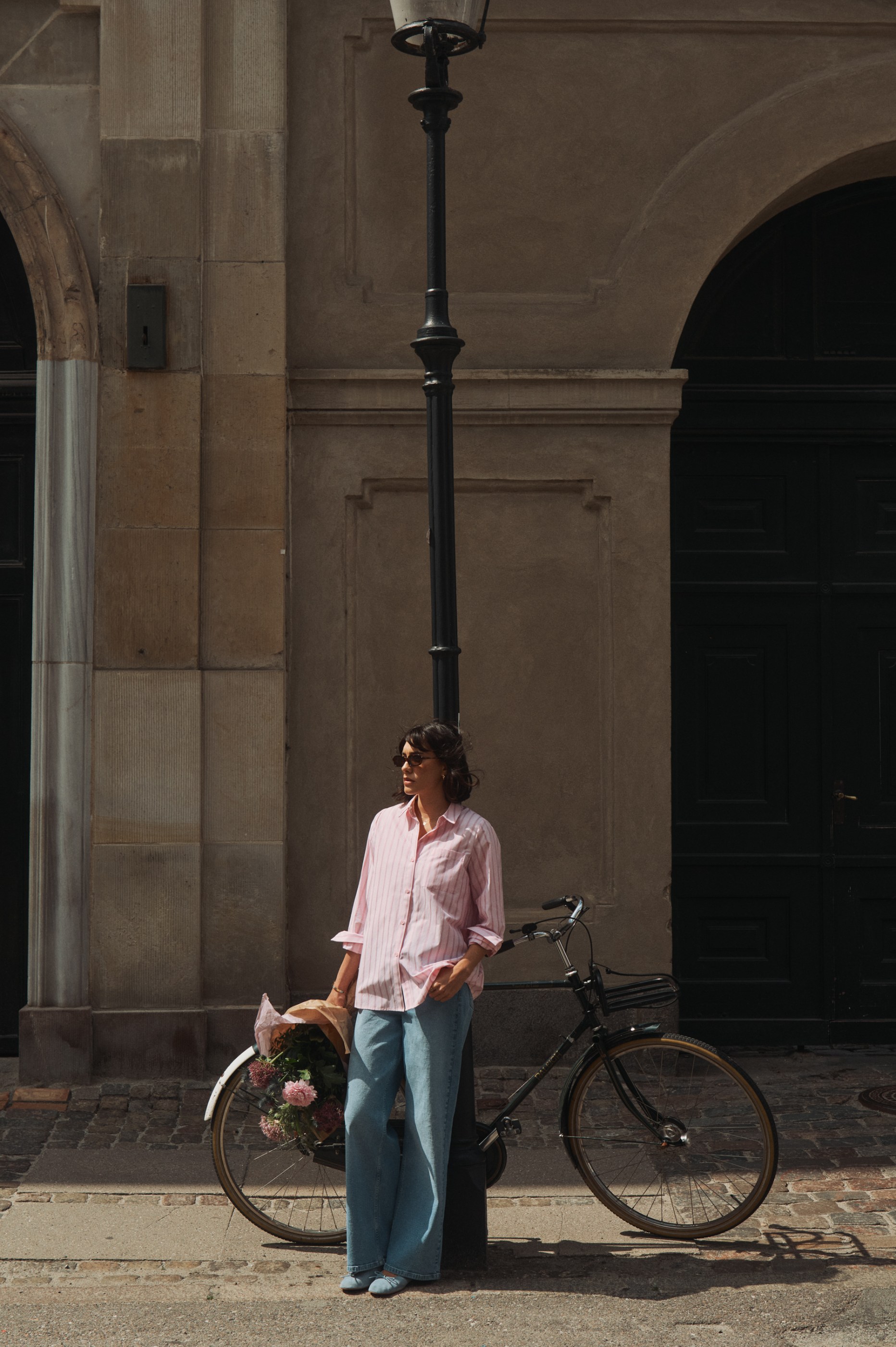 Woman in pink shirt and jeans leans on lamppost with bicycle and flowers on a sunny street.