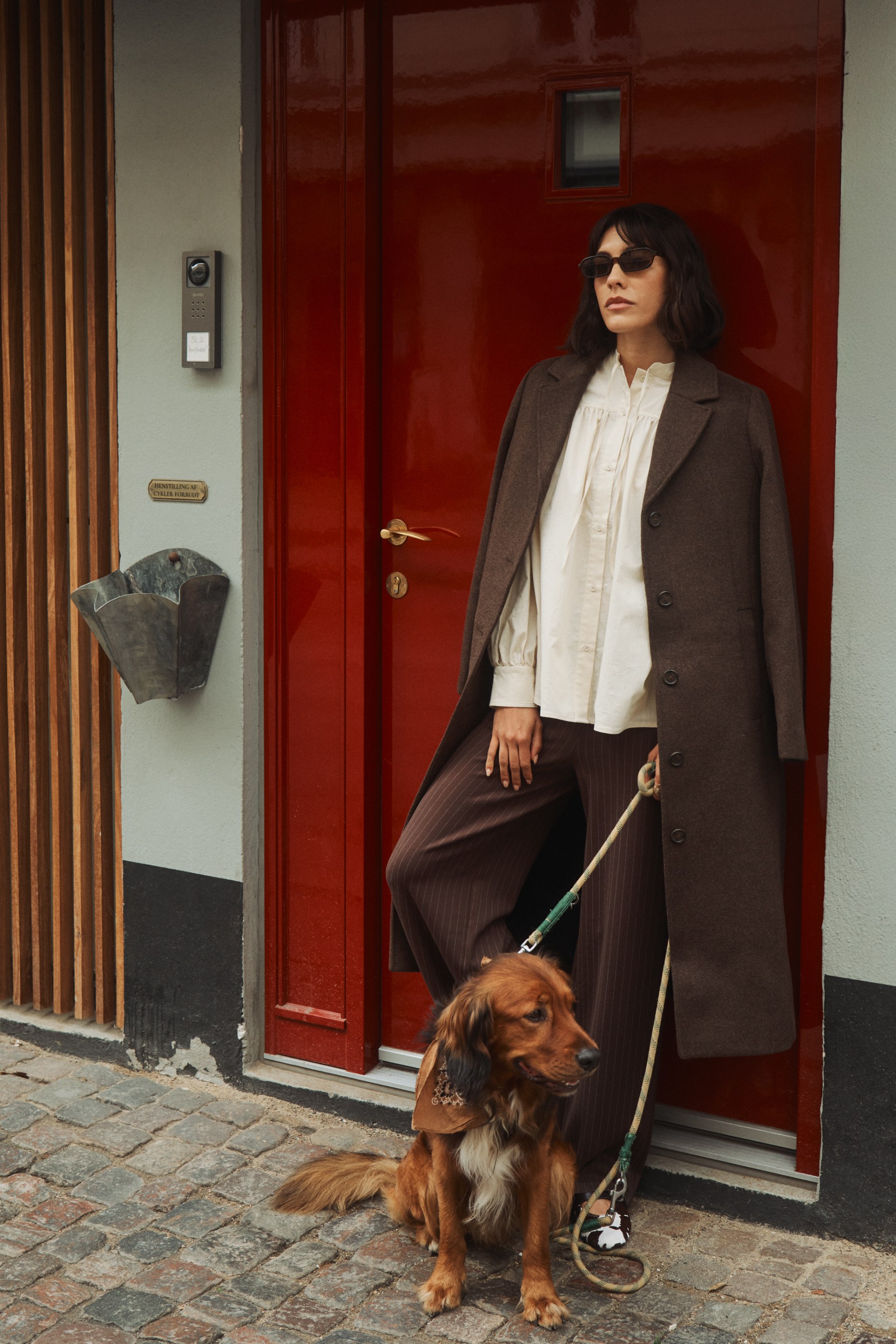 Woman in brown coat and sunglasses stands with a brown dog by a red door.