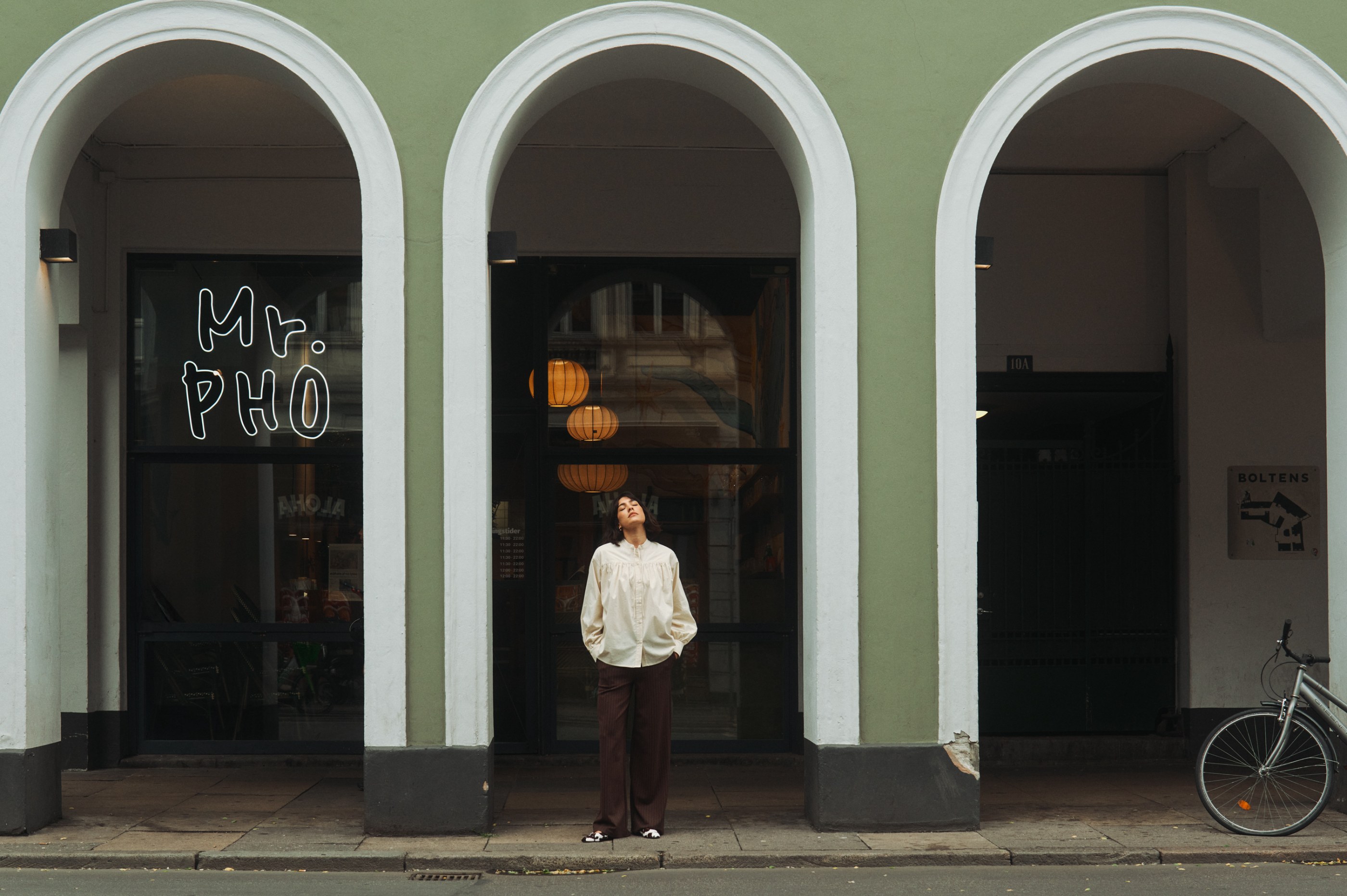 Woman in cream shirt under green arches of a 'Mr. PHO' restaurant, looking up.