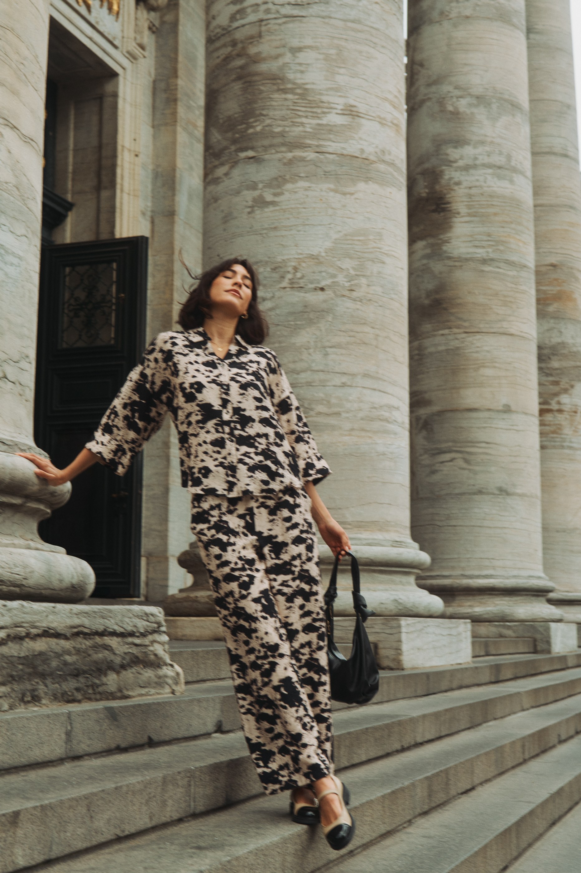 Woman in abstract print outfit, eyes closed, descending grand stone steps and columns.