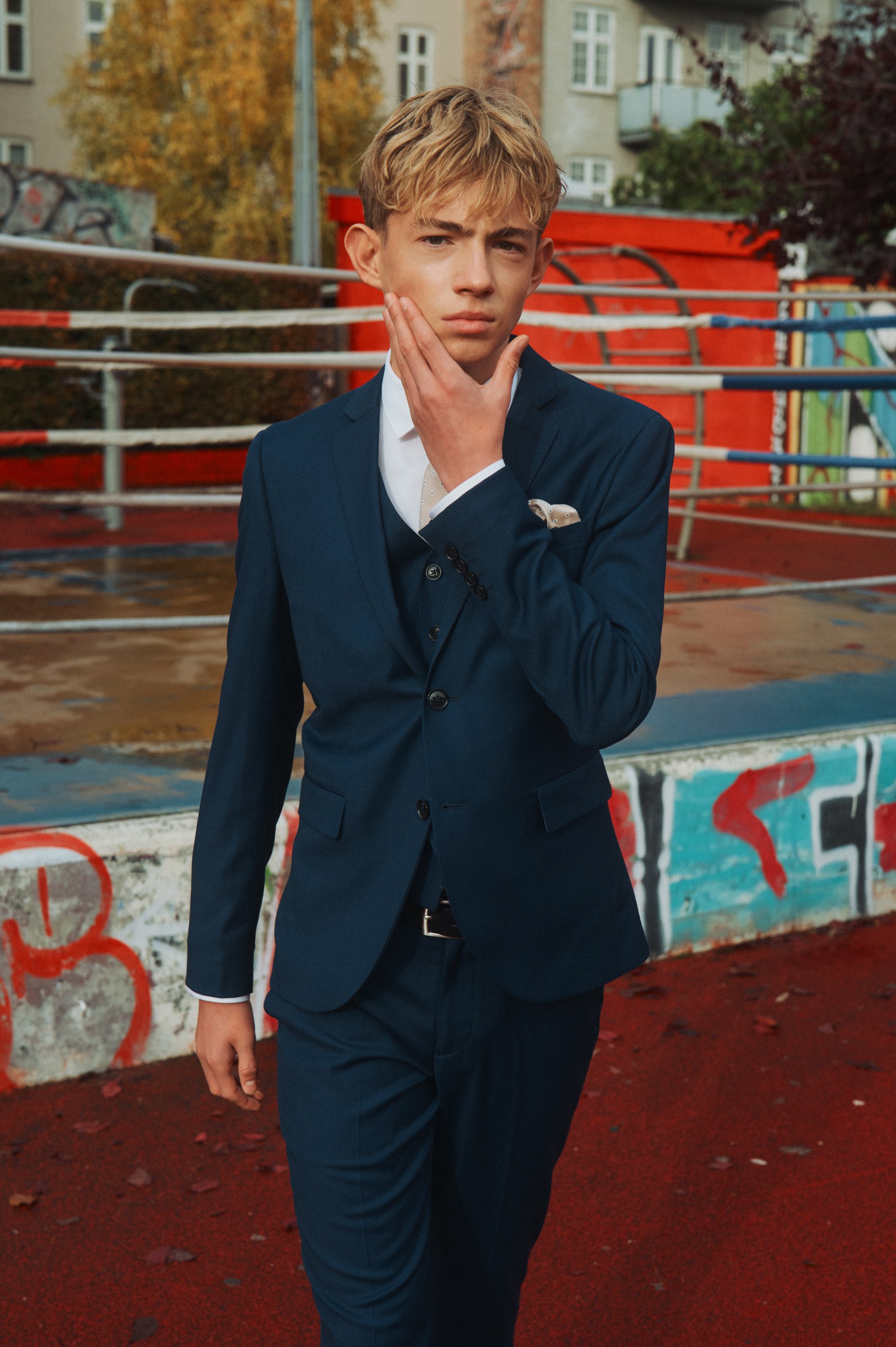 Young man in a navy three-piece suit, hand on chin, in an urban outdoor setting.