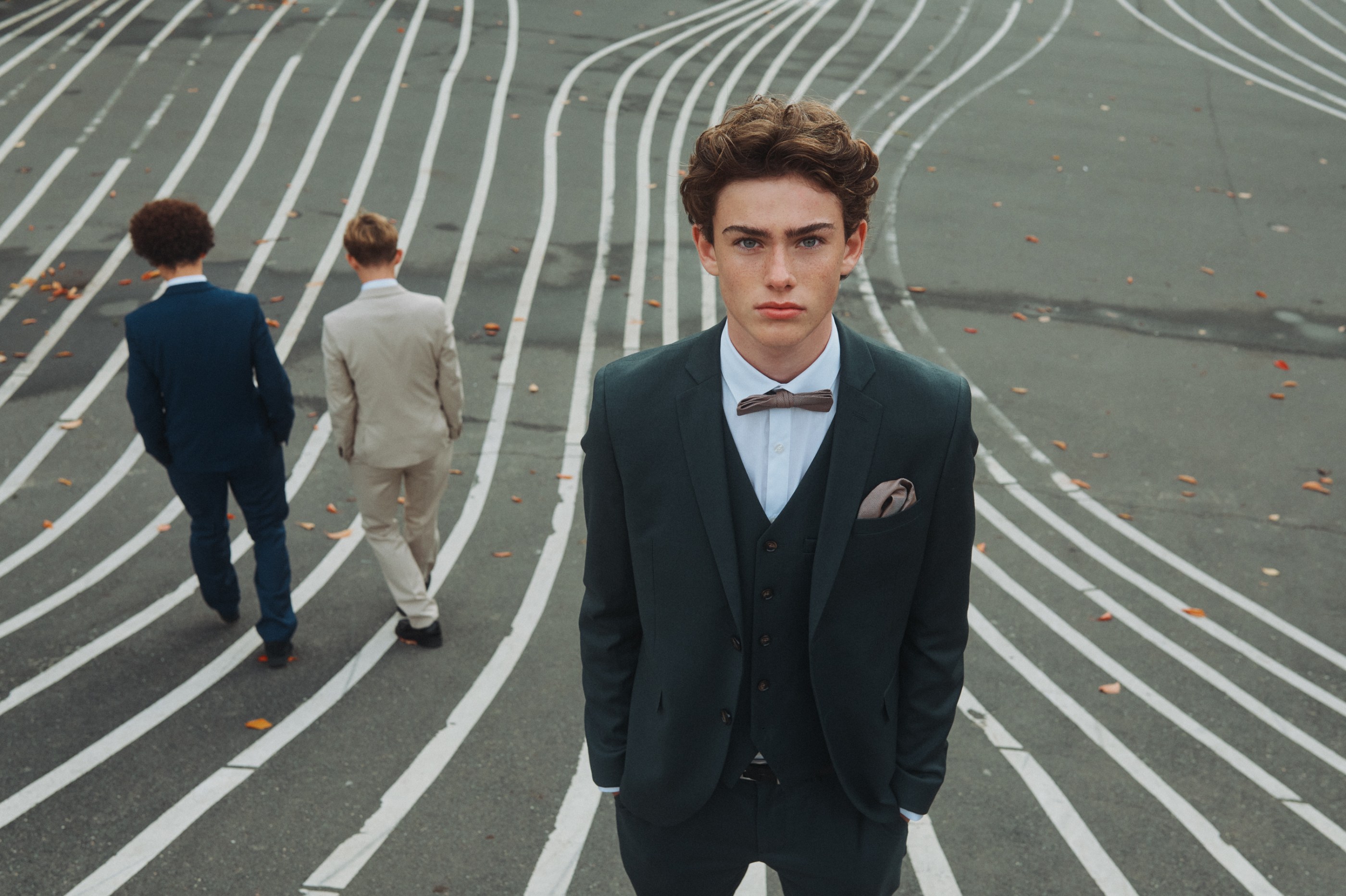 Three young men in suits on a lined outdoor path, one looking directly at camera.