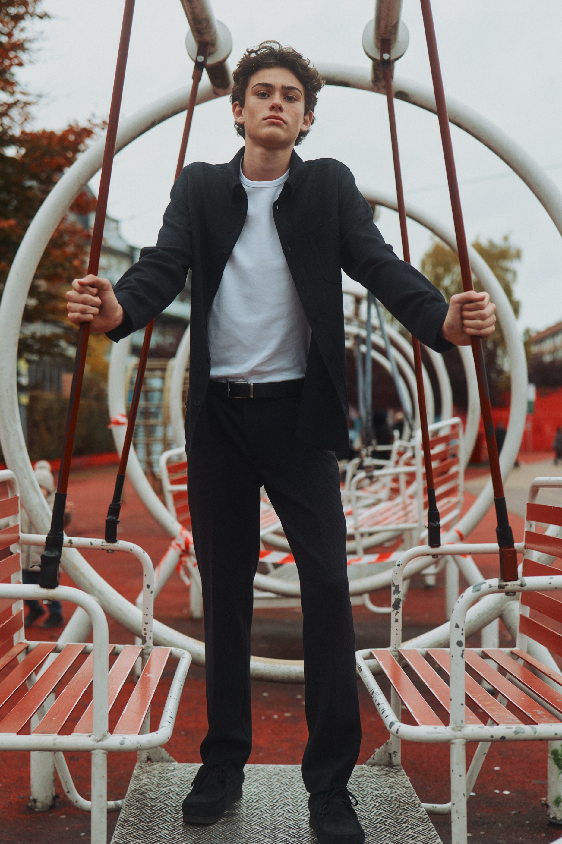 Young man in black jacket and white t-shirt stands on playground swing structure.