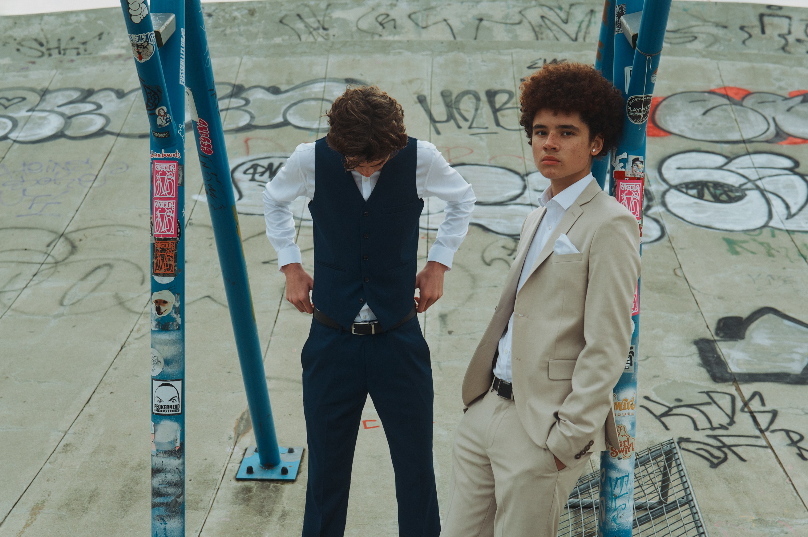 Two young men in formal wear pose among graffiti and blue poles.