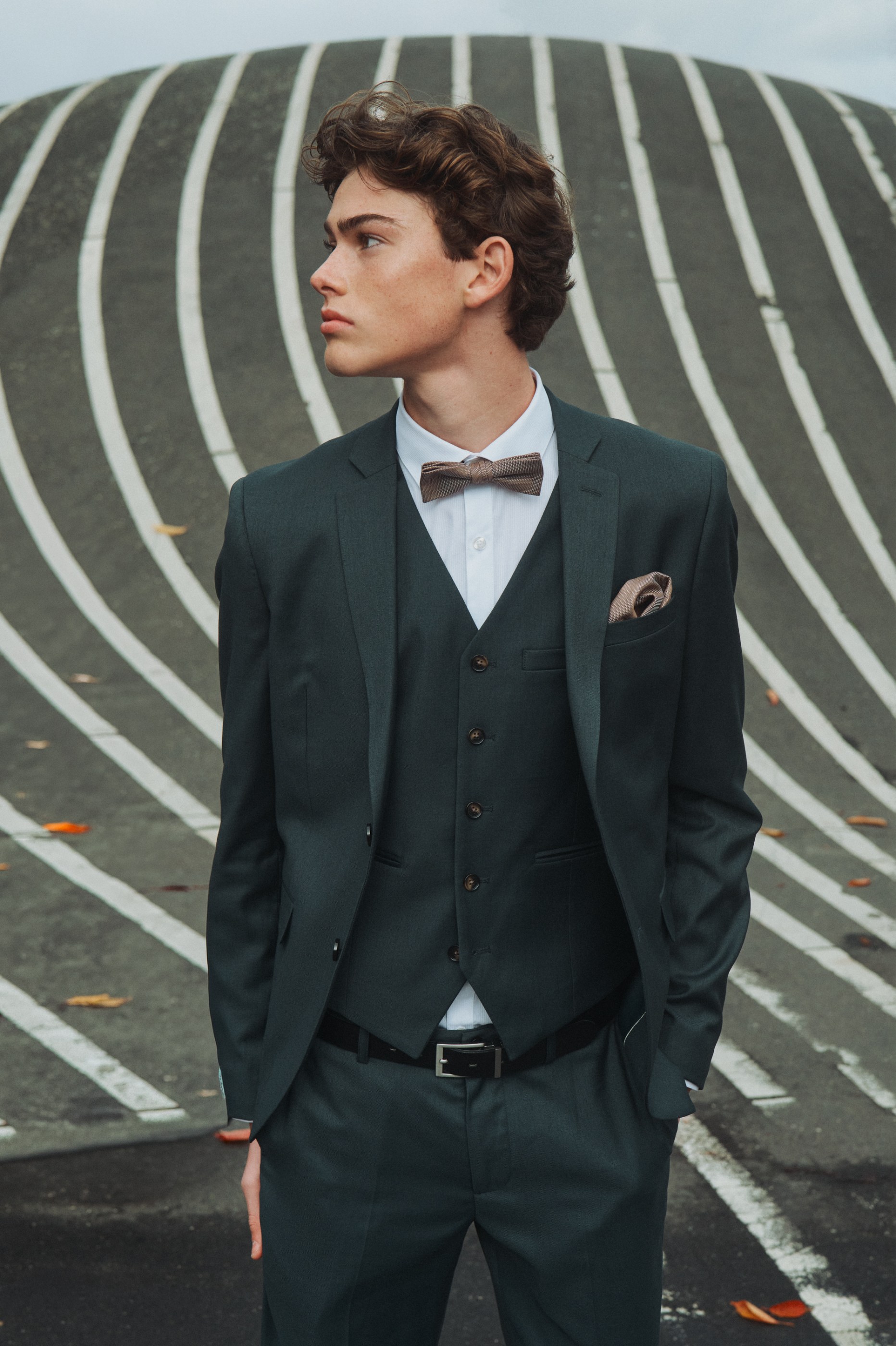 Young man in dark green suit and brown bow tie looks left, striped background.