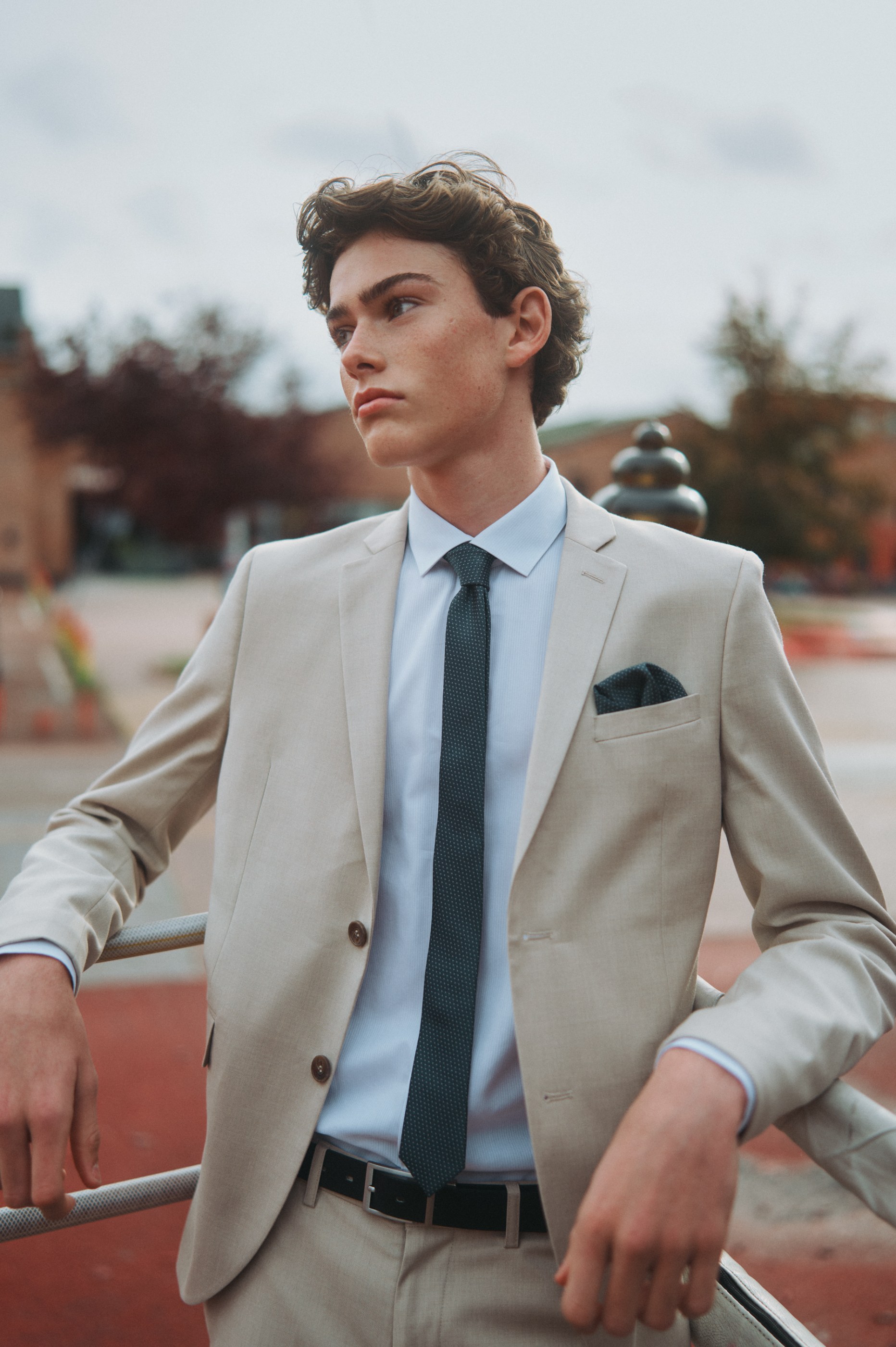 Curly-haired young man in a light tan suit and dark tie leaning on an outdoor railing.