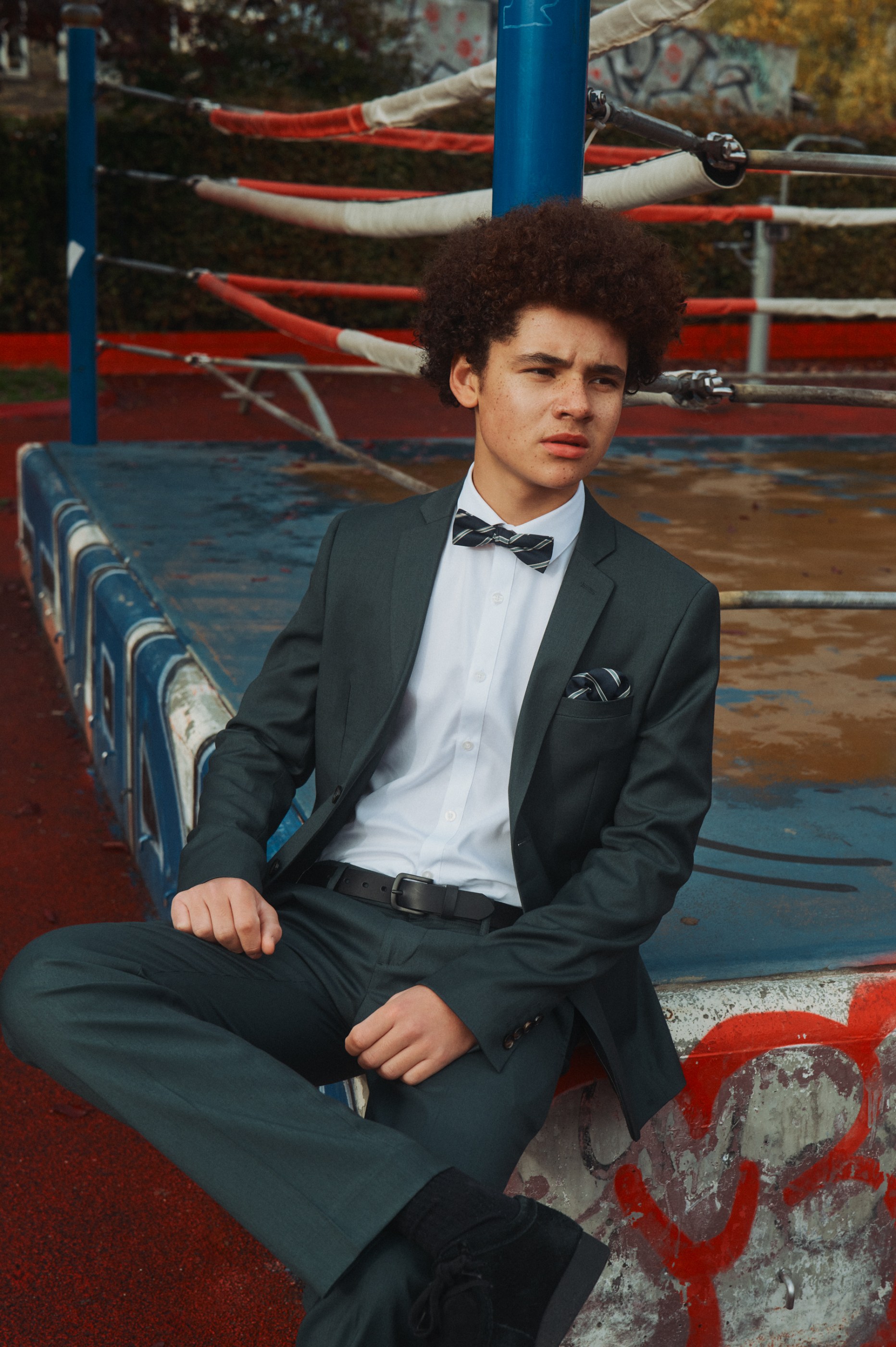 A young man in a dark suit and striped bow tie sits on the edge of a boxing ring.