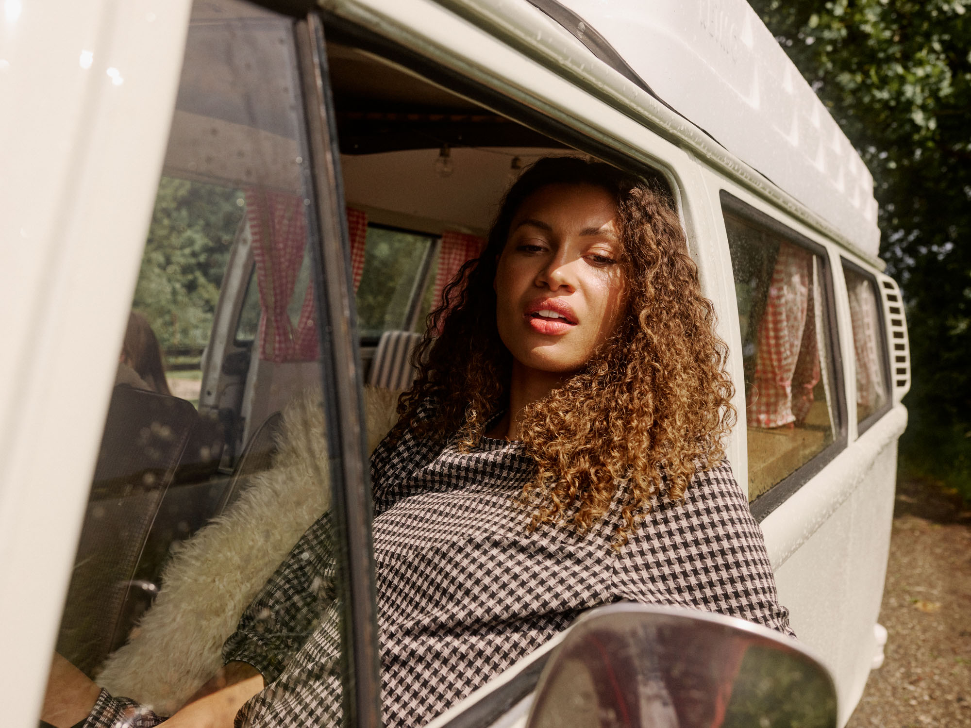 Curly-haired woman in a checked top looking out of a beige camper van window.