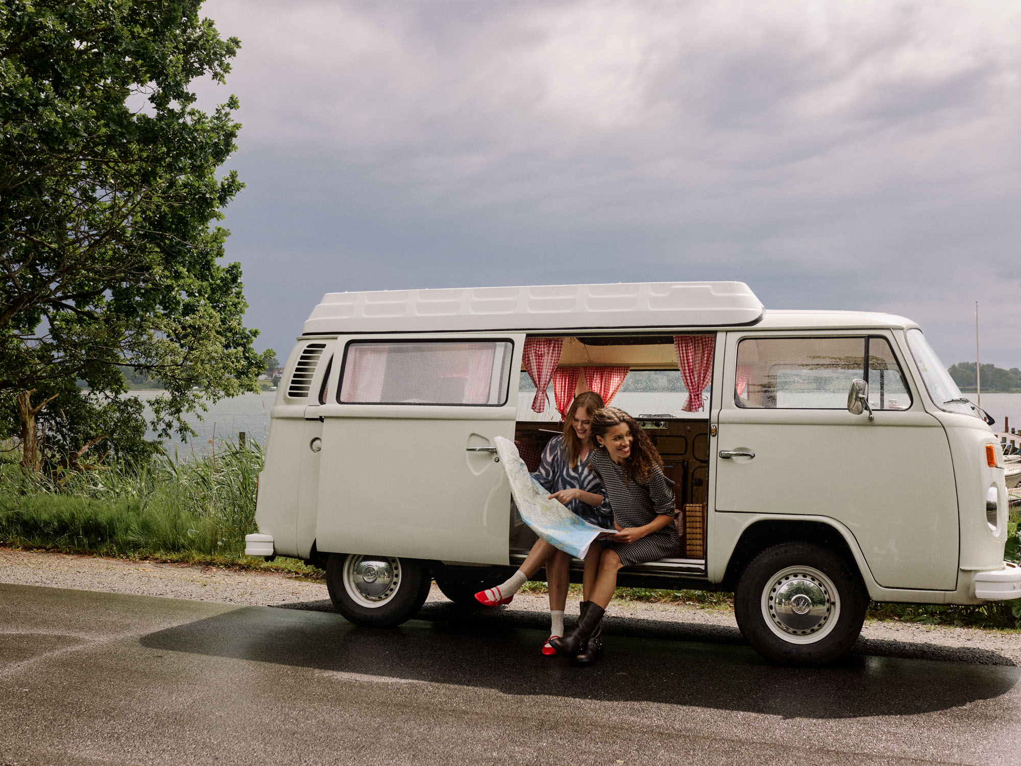Two women looking at a map while sitting in a vintage camper van by a lake.