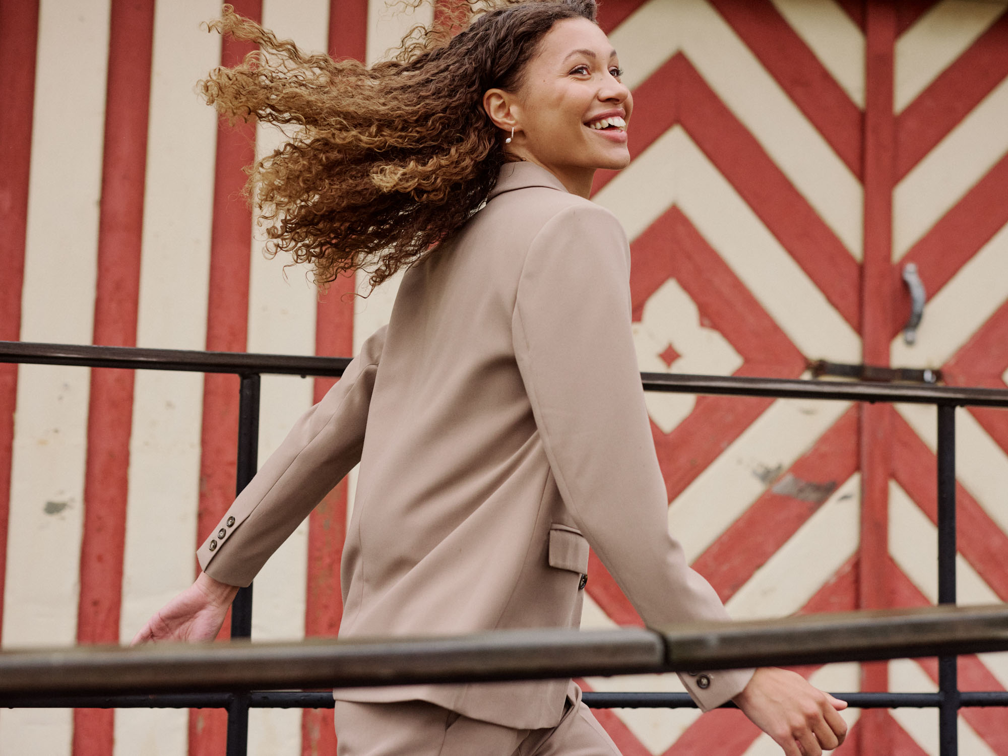 Joyful woman in beige blazer with curly hair blowing, smiling against red and cream patterned wall.