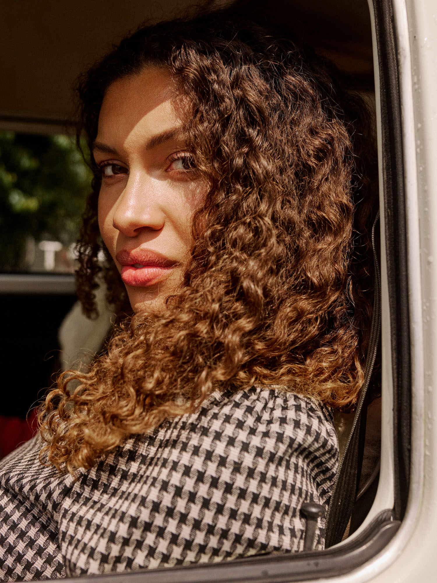 Woman with curly hair and houndstooth top looking out a car window, sunlight on face.