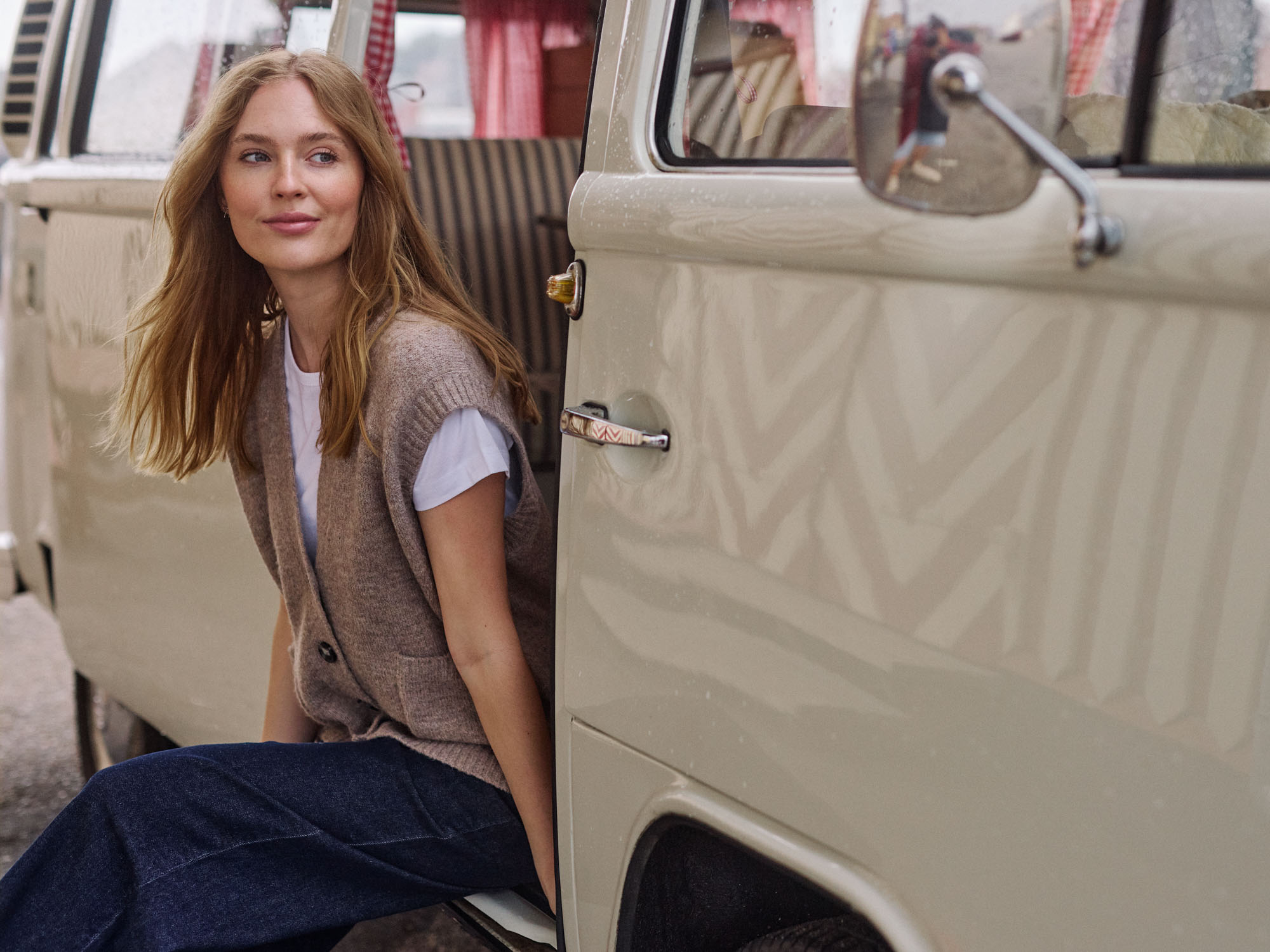 Smiling woman in knit vest, jeans, sitting in a cream vintage camper van doorway.