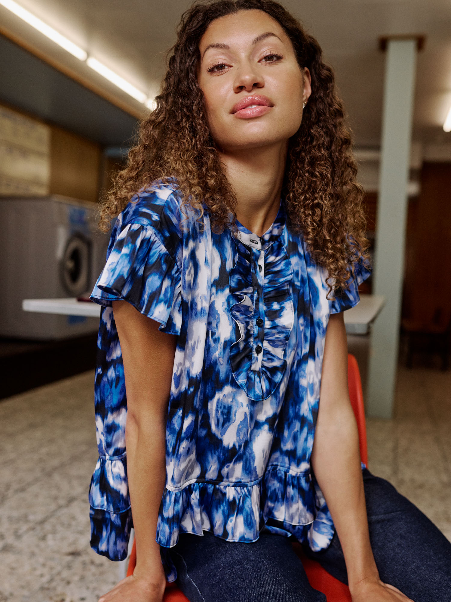 Woman with curly hair in blue and white tie-dye ruffled top sitting in a laundromat.