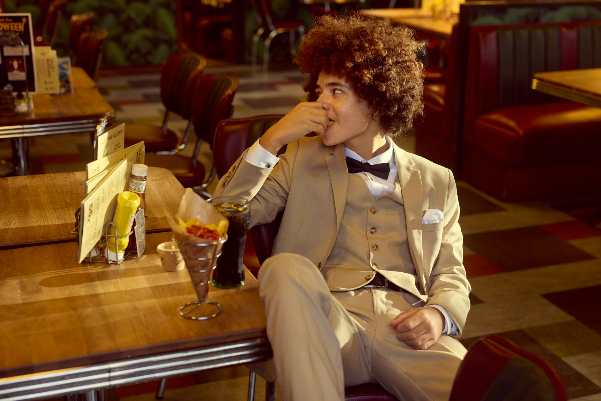 Person with curly hair in a tan suit and bow tie eating at a retro diner.