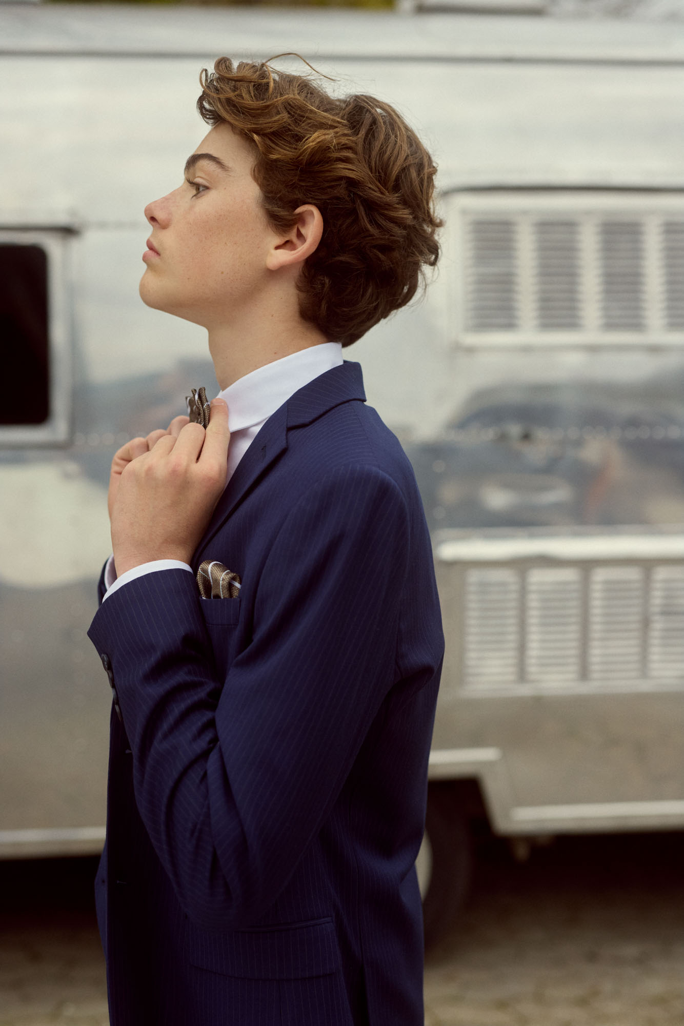 Young man in a navy pinstripe suit adjusting his bow tie, against a silver trailer.