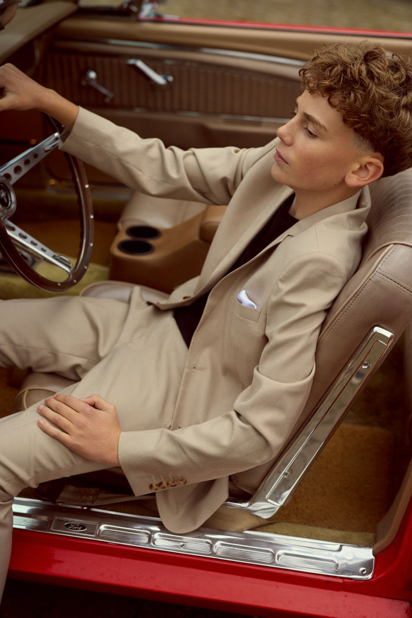 Young man in a beige suit sitting in a red classic car's driver's seat.