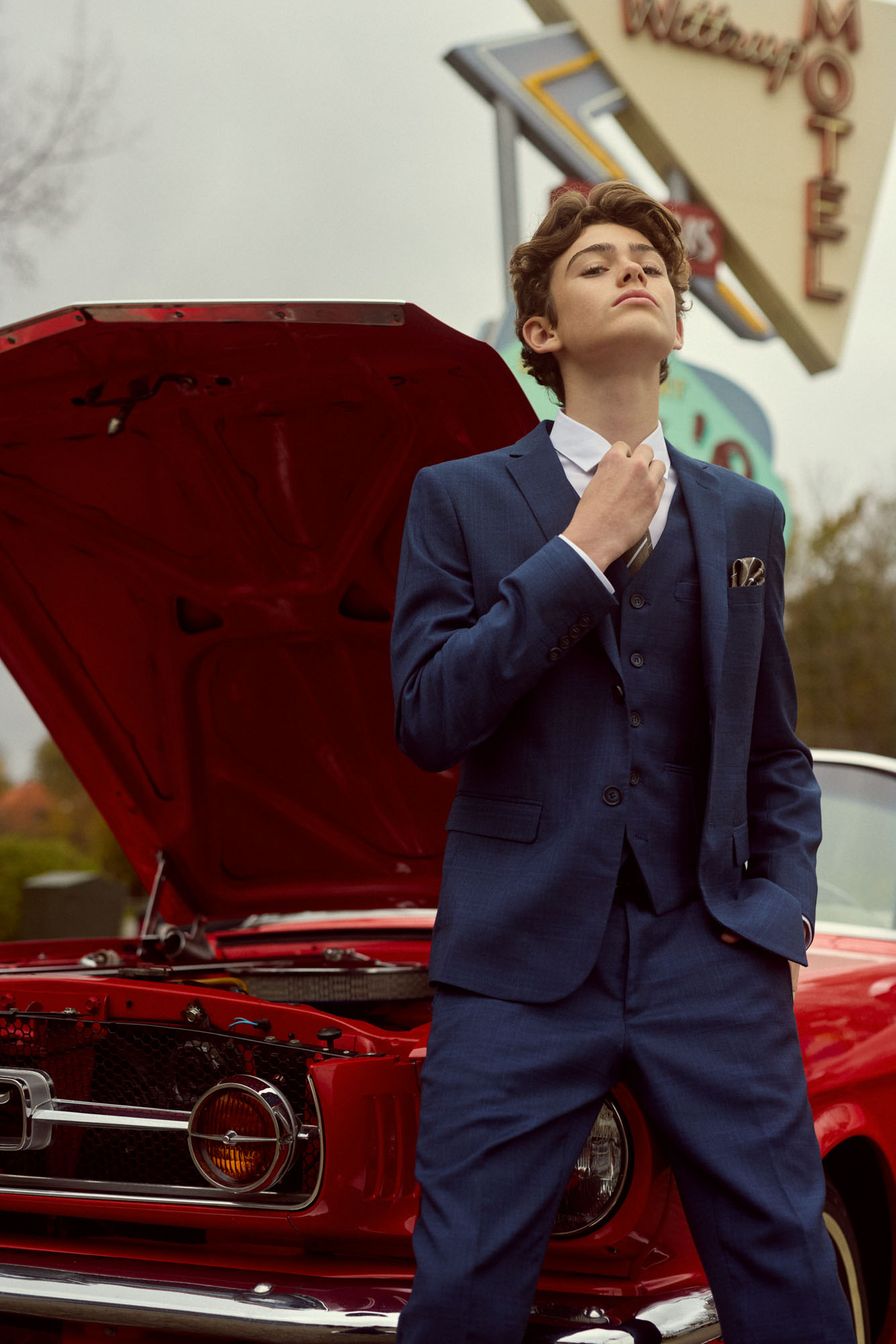 Confident young man in blue suit adjusts tie by classic red car and vintage motel sign.