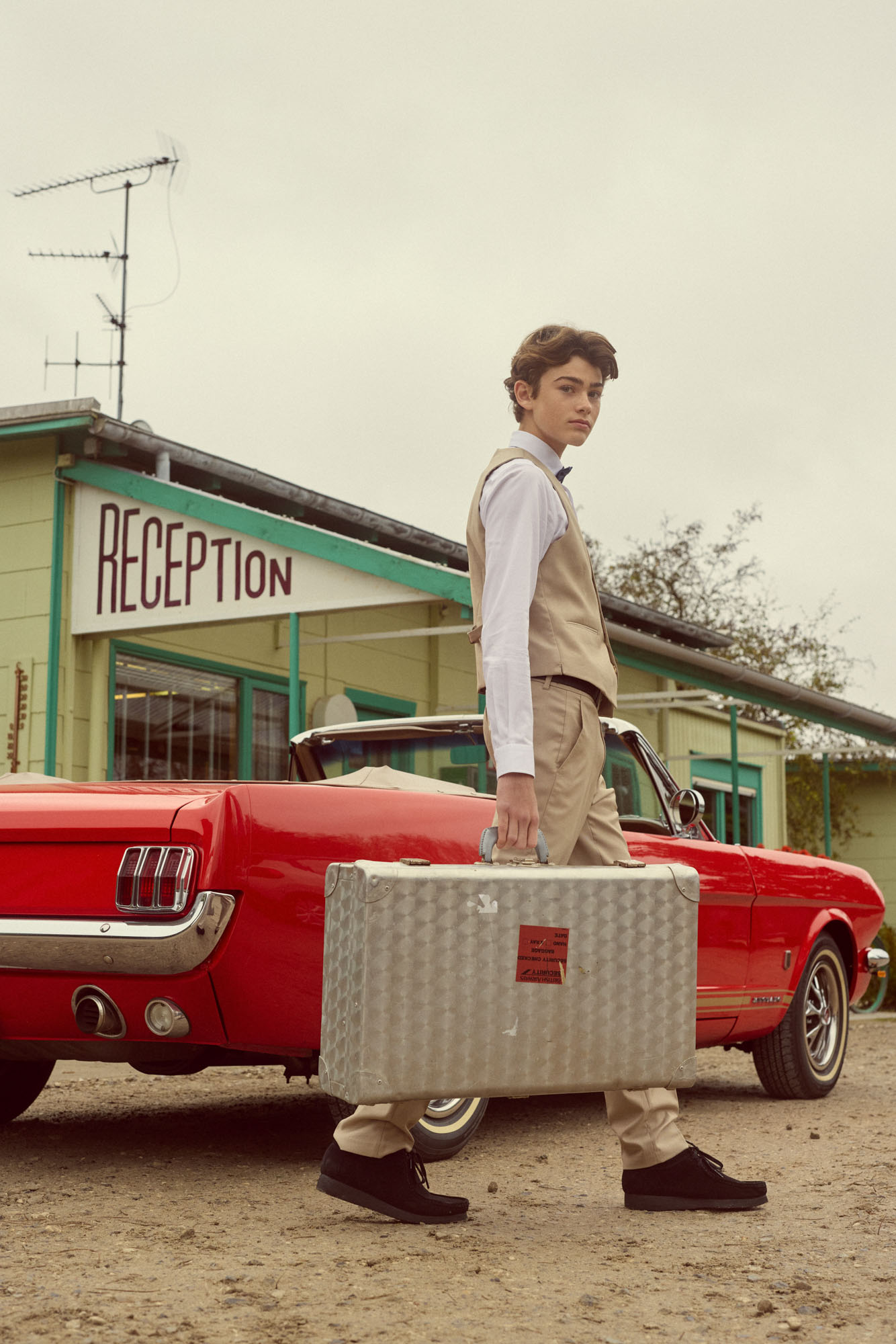 Young man in beige outfit with briefcase walks past red classic car and "RECEPTION" building.