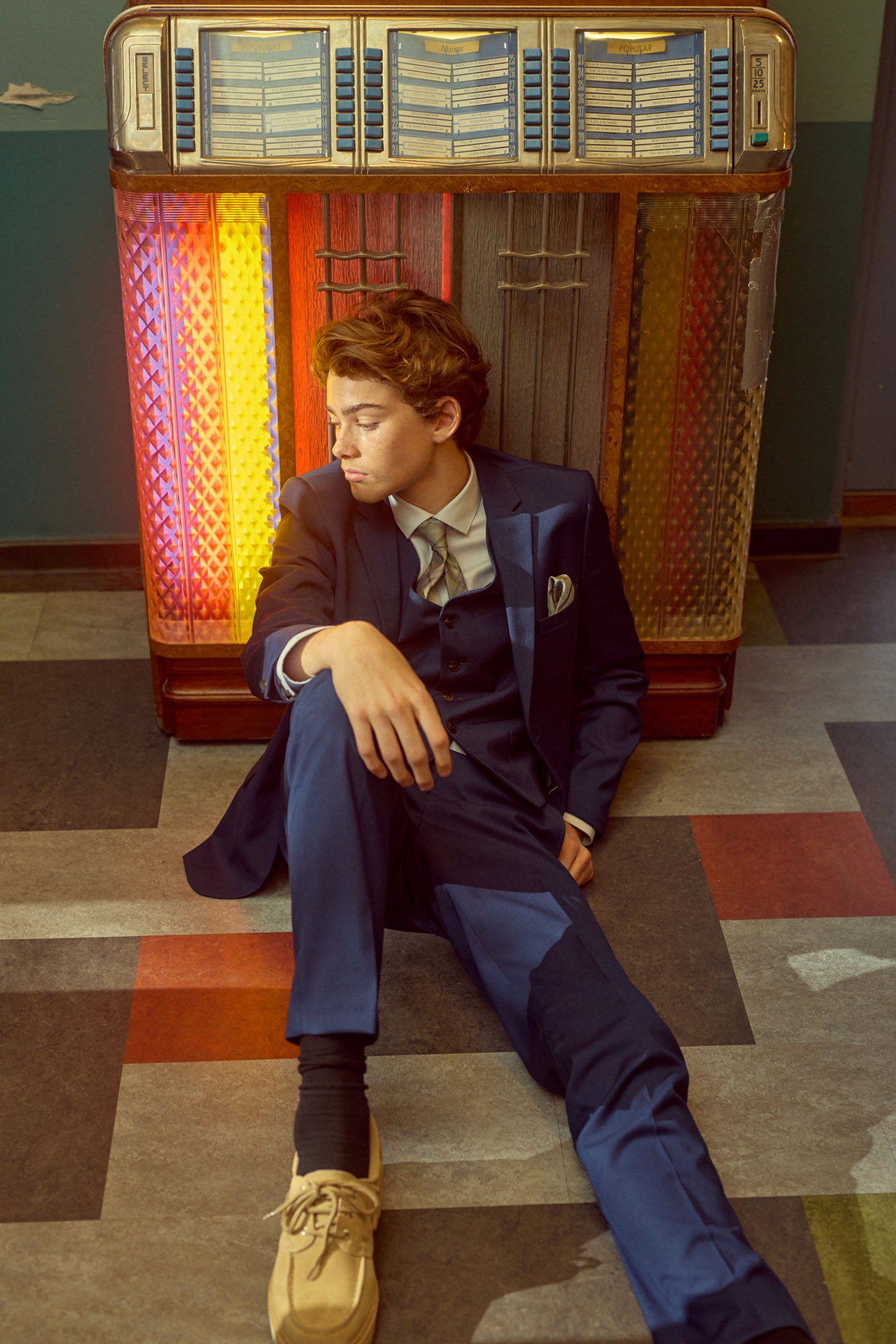 Pensive young man in a blue suit sits on a tiled floor by a glowing vintage jukebox.