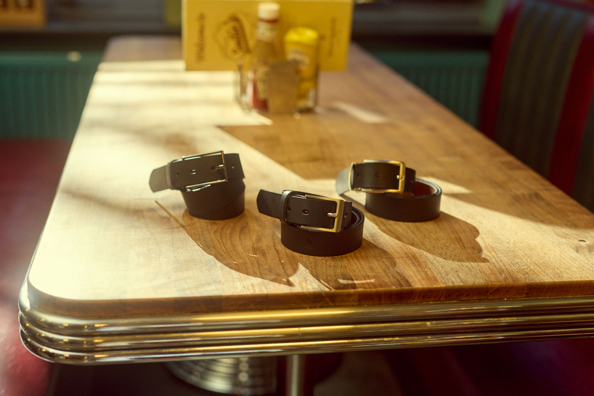Three black leather belts with metal buckles on a sunny wooden diner table.