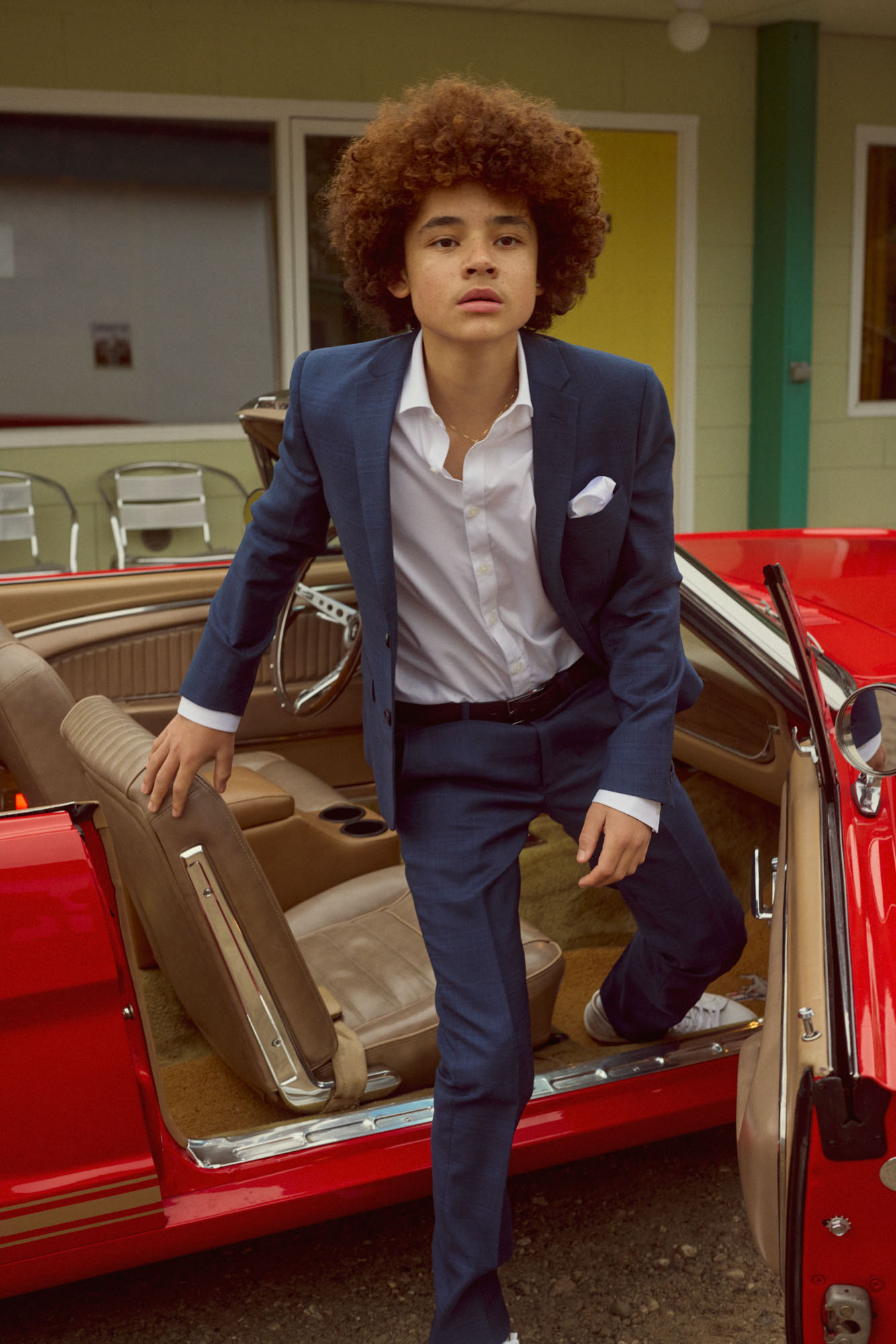 Young person with curly hair getting out of a red vintage convertible wearing a navy suit.